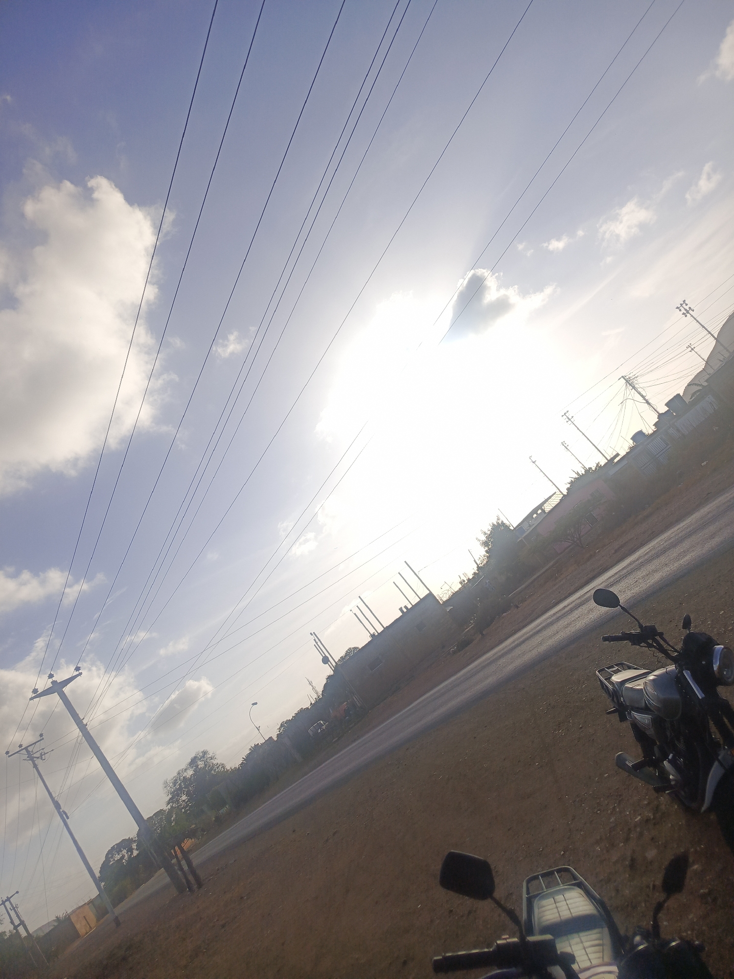 The image captures a street scene in Juan Griego, Venezuela under a bright, sunlit sky with scattered clouds. Power lines stretch across the upper portion of the frame. A paved road runs diagonally through the image, with buildings and vegetation visible along the roadside. Two motorcycles are prominently featured in the foreground. The lighting suggests it is daytime.