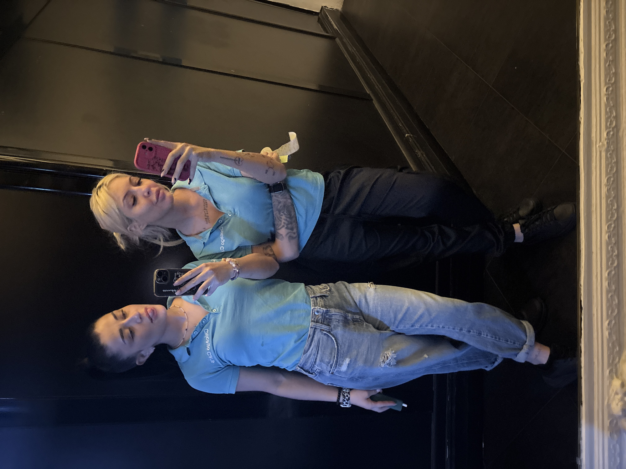 Two women are taking a selfie in a mirrored elevator. The woman on top has blonde hair and is wearing black pants and a light blue collared shirt. She is holding a pink phone. The woman below has dark hair and is wearing ripped jeans and a light blue collared shirt. She is holding a black phone. The elevator has black walls and flooring. The location is likely not in Plovdiv, Bulgaria, as the shirts worn by the two women have "Chicago CSLA" printed on them.