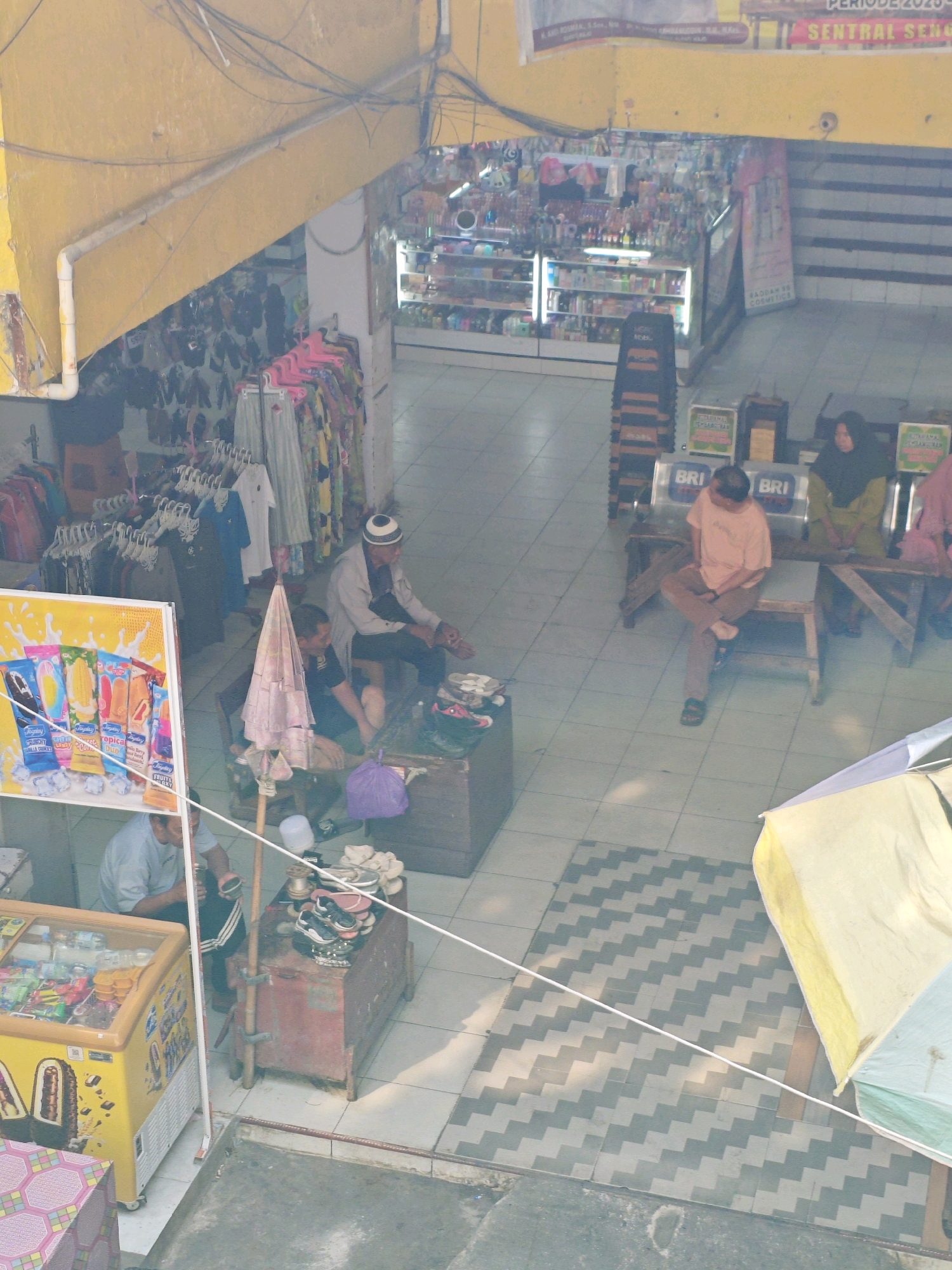 The image captures a scene within a marketplace in Teddaopu, Indonesia. In the foreground, a yellow refrigerated display case filled with frozen treats and ice cream is visible. To the left, a person wearing a light blue shirt and dark pants is seated, holding what appears to be a dark object. In the middle ground, a wooden bench is occupied by three individuals. One person, wearing a light pink shirt and brown pants, sits with their legs crossed, looking downwards. To their right, a person in a dark shirt and headscarf sits with their arms resting on their lap. Further to the right, another person wearing a light brown shirt and dark pants is seated. Adjacent to the bench, a wooden structure with items on it, possibly for sale or storage, is present. A purple plastic bag rests on the floor near this structure. To the left of the bench, an older man wearing a white cap and a light grey outer garment is seated on a wooden stool. He is looking down at something in front of him. Near him, a second person in a dark shirt is also seated. A pink umbrella leans against a wooden pole. In the background, numerous stalls and shelves are stocked with a variety of goods, including clothing and cosmetic products. The floor is tiled in a checkered pattern in some areas and a zigzag pattern in others. Overhead, electrical wires are strung across the ceiling. The overall atmosphere suggests a busy commercial environment.