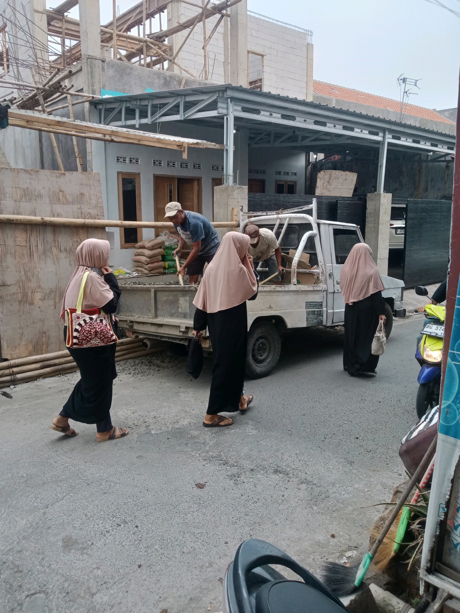 In Grendeng, Indonesia, a construction site is active. A man in a blue shirt and a beige cap is shoveling aggregate from a white pickup truck into a cement mixer. Three women, two in black abayas and pink hijabs, and one in a black abaya and pink hijab, are walking or standing nearby. The woman on the far left carries a red and white patterned tote bag. The woman in the center walks with her head down, and the woman on the far right carries a light-colored woven bag. In the background, a building is under construction with exposed beams and scaffolding. A yellow motorcycle is parked to the right of the truck. The scene appears to be on a paved street in front of the construction site, with bamboo poles and piles of cement bags visible.