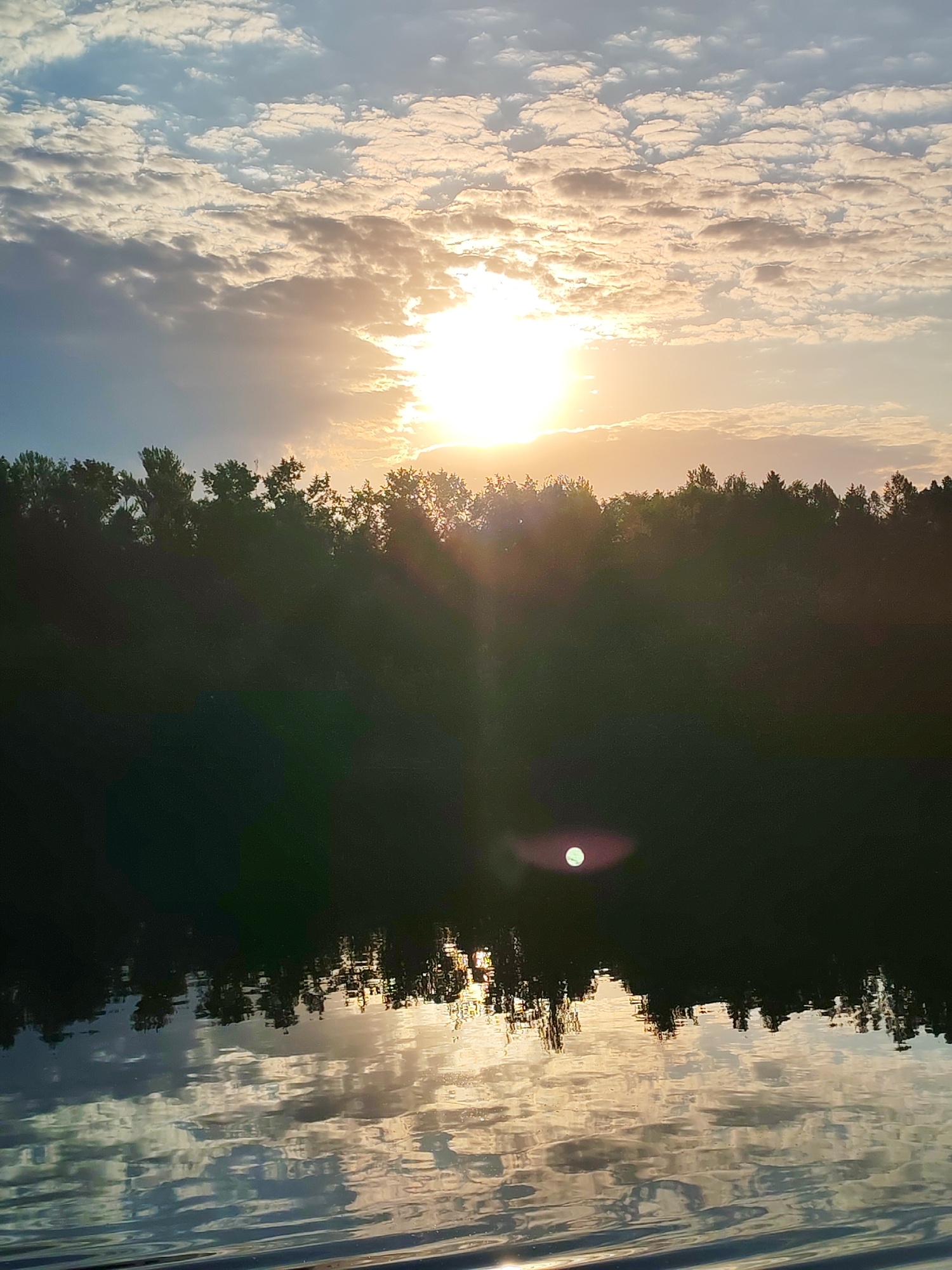 The image depicts a landscape at sunset. The sky is filled with altocumulus clouds, some illuminated by the setting sun. The sun itself is positioned above a treeline, casting rays of light downwards. A body of water occupies the lower portion of the frame, reflecting the sky and treeline. The reflections are distorted by ripples on the water's surface. The treeline appears dark, silhouetted against the bright sky. The location is Letchik-Ispytatel', Russia.
