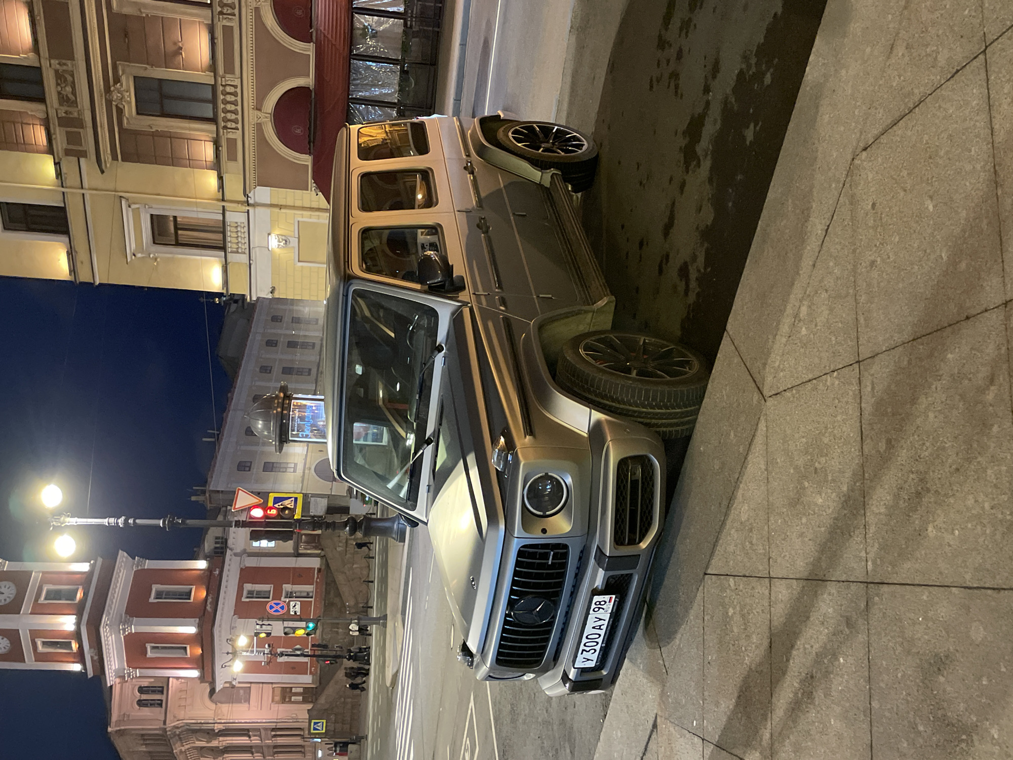 The image shows a gray Mercedes-Benz G-Class parked on a street in Saint Petersburg, Russia. The license plate number is V300AY98. The vehicle is parked on the right side of the street, near the curb of the sidewalk. In the background, the Nevsky Prospekt is visible. Buildings on the street are illuminated. There is a traffic light with the green light on, and people are seen walking on the crosswalk. The sky is dark. The clock tower building is visible in the background.