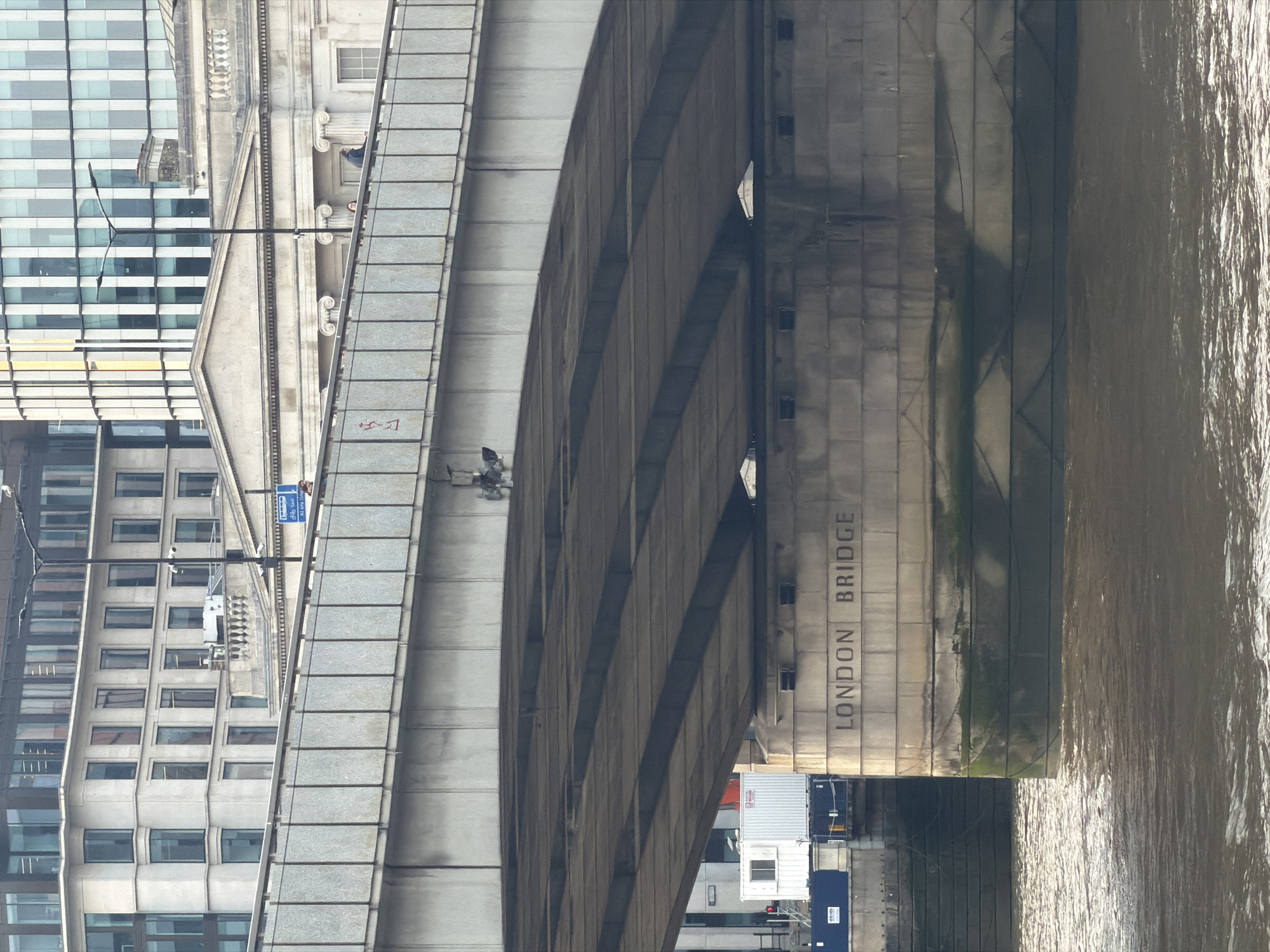 The image depicts London Bridge in London, United Kingdom, viewed from an elevated position. The bridge is constructed of grey concrete, with the words "LONDON BRIDGE" vertically inscribed on one of the structural supports. The bridge spans a body of water that reflects light, creating a textured surface. Adjacent to the bridge on the left is a series of buildings with multiple stories and many windows, some of which are obscured by shadows. On the bridge structure, a few people can be seen walking. A sign is visible near the walkway, suggesting a route for pedestrians and vehicles. The overall scene is a mix of urban infrastructure and natural elements.