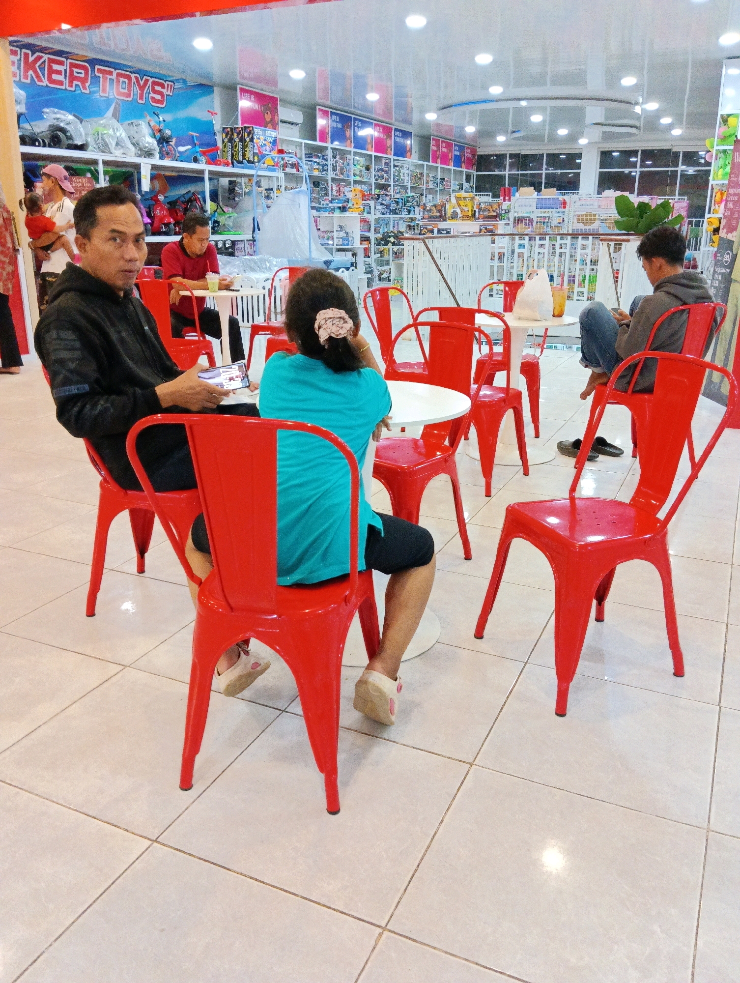 The image depicts a brightly lit interior, likely a toy store given the "KER TOYS" signage visible. The flooring is tiled in a light beige color, and the scene appears to be a seating area within the store.
In the foreground, a man in a black hoodie is seated on a red metal chair, looking towards the camera and holding a smartphone. To his right, a person wearing a teal shirt and black shorts sits with their back to the camera, also on a red metal chair. Their hair is tied up with a pink accessory.
Further back, another man in a red shirt is seated at a white table, looking down at a drink. A child in a red outfit is being held by an unseen person to the left. In the background, shelves are filled with various items, predominantly toys, and displays of products are visible throughout the store. Another person in a gray hoodie is seated on a red chair to the far right, looking at their phone. The environment suggests a commercial setting, possibly a cafe or rest area within a retail space. The overall impression is of a casual gathering or break within a busy shopping environment.