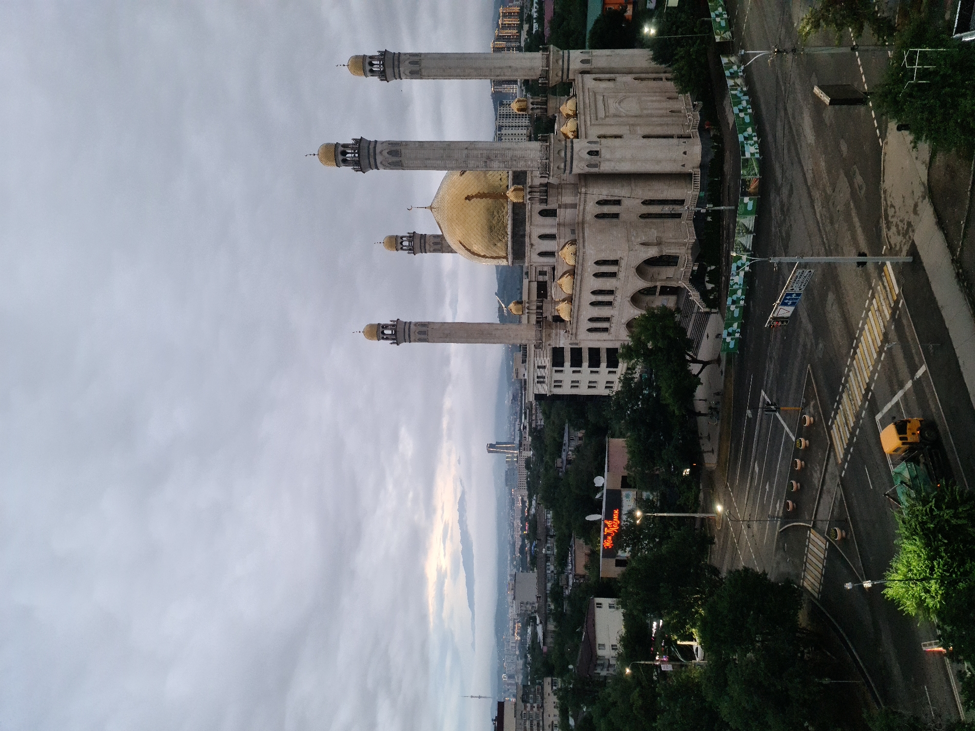 The image depicts the large Central Mosque of Almaty, Kazakhstan, under a cloudy sky. The mosque is a prominent structure with a large golden dome and four tall minarets, also topped with golden finials. The building is constructed of light-colored stone and features arched windows and doorways.
In the foreground, a street intersection with crosswalks is visible. A yellow garbage truck is making a turn at the intersection. The surrounding area consists of low-rise buildings, trees, and streetlights. In the background, a cityscape stretches towards hazy mountains. A tall, modern skyscraper and a radio tower are also visible in the distance. The overall atmosphere is overcast and somewhat muted, suggesting either early morning or a heavily clouded day.