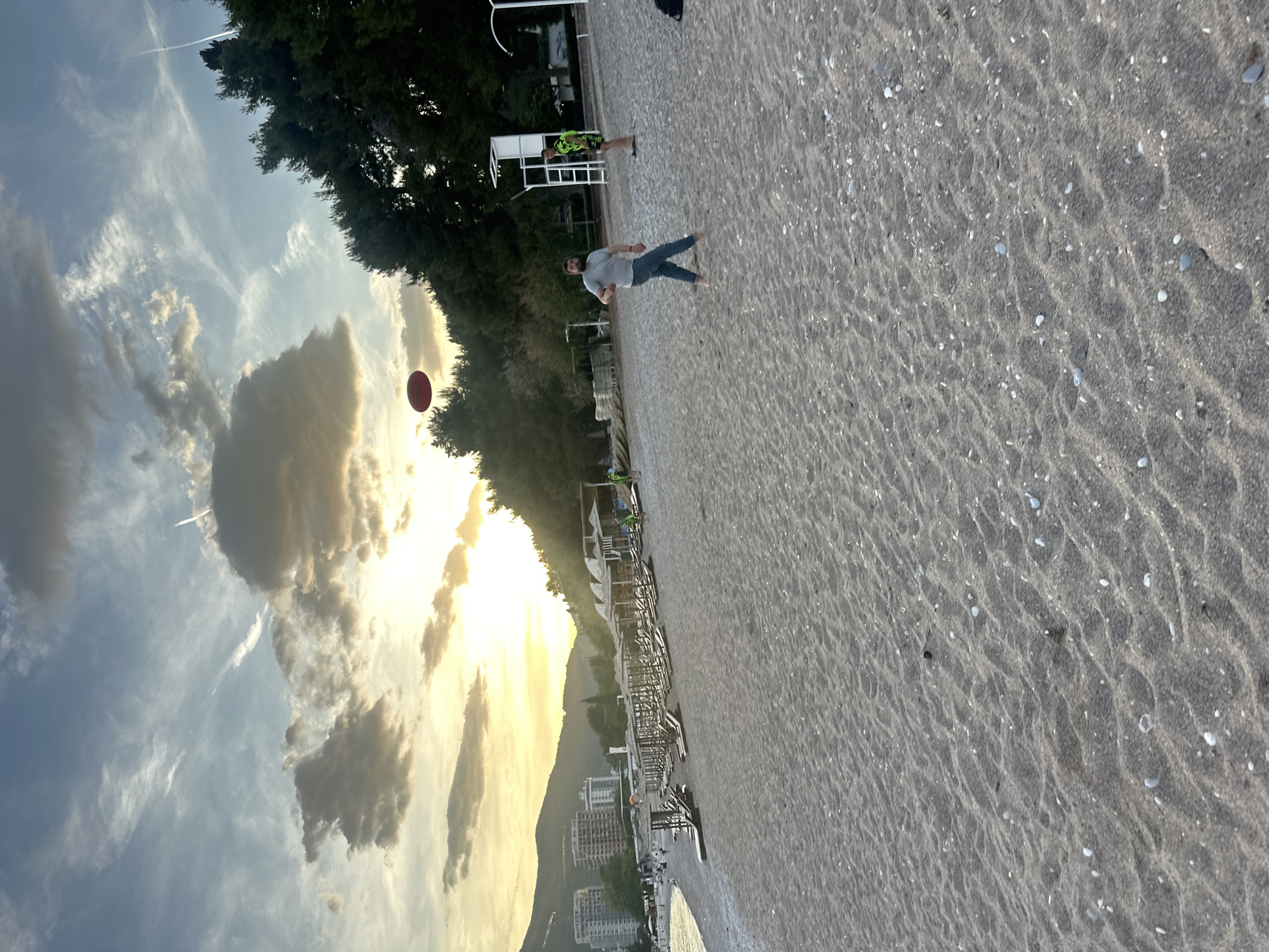 The image captures a scene at Mogren Beach in Budva, Montenegro, looking westward toward the setting sun. The beach consists of coarse, gray sand with scattered shells. A man stands barefoot on the sand, looking upward and right toward a floating red ball or Frisbee against the backdrop of the sun and dramatic cloud formations. In the background, there is a row of beach umbrellas and lounge chairs extending along the shoreline. The cityscape of Budva is visible at the base of a mountain range in the distance. A lifeguard stand is present near the trees. Another person in a green shirt stands away in the background. The sky is filled with streaky clouds illuminated by the sunset.