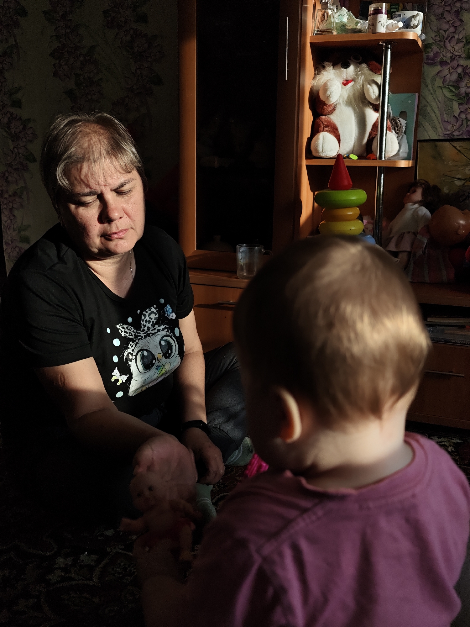 The image shows an interior scene. An adult woman with short, graying hair sits on the floor wearing a black t-shirt with a cartoon owl print. She holds a small doll in her hands. A toddler, with short, light-colored hair, stands facing away from the camera, wearing a pink t-shirt.
In the background is a wooden cabinet or shelving unit filled with various items including stuffed animals, dolls, and a stacking toy. Wallpaper with a floral pattern is visible behind the cabinet. The lighting is mixed, with portions of the scene in shadow and others in direct sunlight.
The location is indicated as Уральское, Russia.