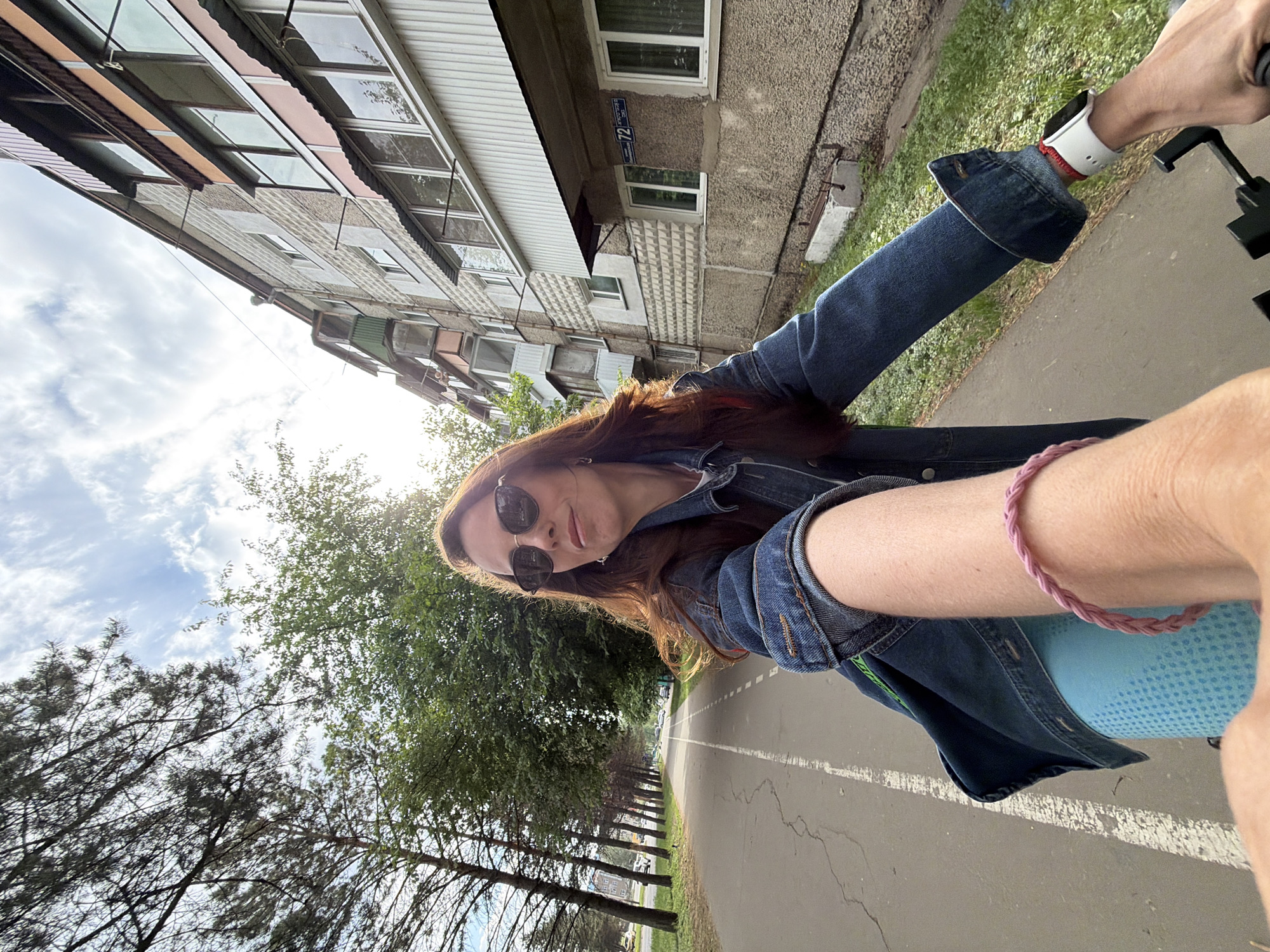 The image shows a woman taking a selfie while riding a bicycle on a paved path lined with trees. The path appears to be a dedicated bike lane, indicated by a white line. She is wearing a denim jacket, sunglasses, and a watch. A pink bracelet is visible on her arm. A building is visible to the left, identified by the street number "72". This indicates the location is Birobidzhan, Russia. The sky is partly cloudy, suggesting it is daytime. The woman is looking directly at the camera.