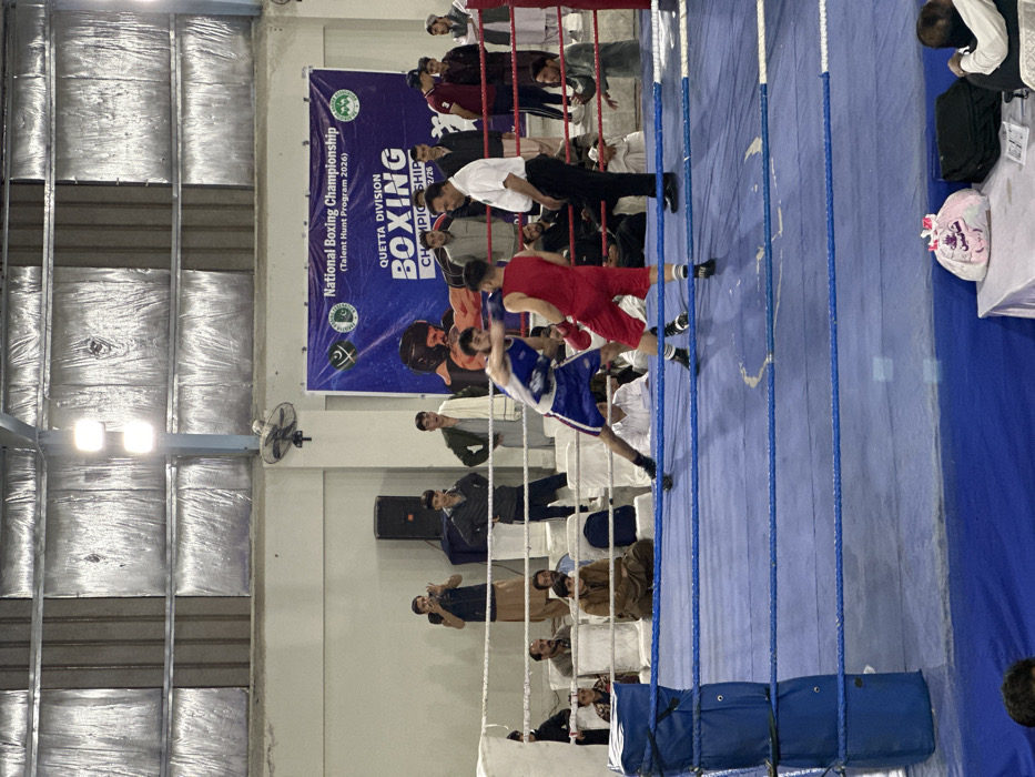The image displays an indoor boxing championship event in Quetta City Tehsil, Pakistan. Two male boxers are actively engaged in a match within a blue-roped boxing ring with blue corner pads. The boxer on the left wears a blue sleeveless top, blue shorts, and red boxing gloves, appearing to throw a left punch. The boxer on the right wears a red sleeveless top, red shorts, and red boxing gloves, positioned defensively. A male referee in a white short-sleeved shirt and black trousers stands between the boxers, observing their actions.
Approximately 25-30 male individuals are present outside the boxing ring, functioning as spectators or event staff. Some are seated on white plastic chairs arranged around the ring, while others stand. One individual on the left side of the ring is recording the event with a mobile phone or camera.
In the background, a large blue banner with white text is affixed to a light-colored wall, featuring the Pakistani flag and other logos. The banner explicitly reads "National Boxing Championship (Talent Hunt Program 2026) QUETTA DIVISION BOXING CHAMPIONSHIP." Above the banner, a wall-mounted fan is visible. To the left of the banner, a dark speaker system is positioned. The venue ceiling features exposed metal trusses and silver insulation panels, with multiple overhead light fixtures illuminating the scene. In the bottom right foreground, a table covered with a white cloth holds a white plastic bag with pink writing and a dark bag.