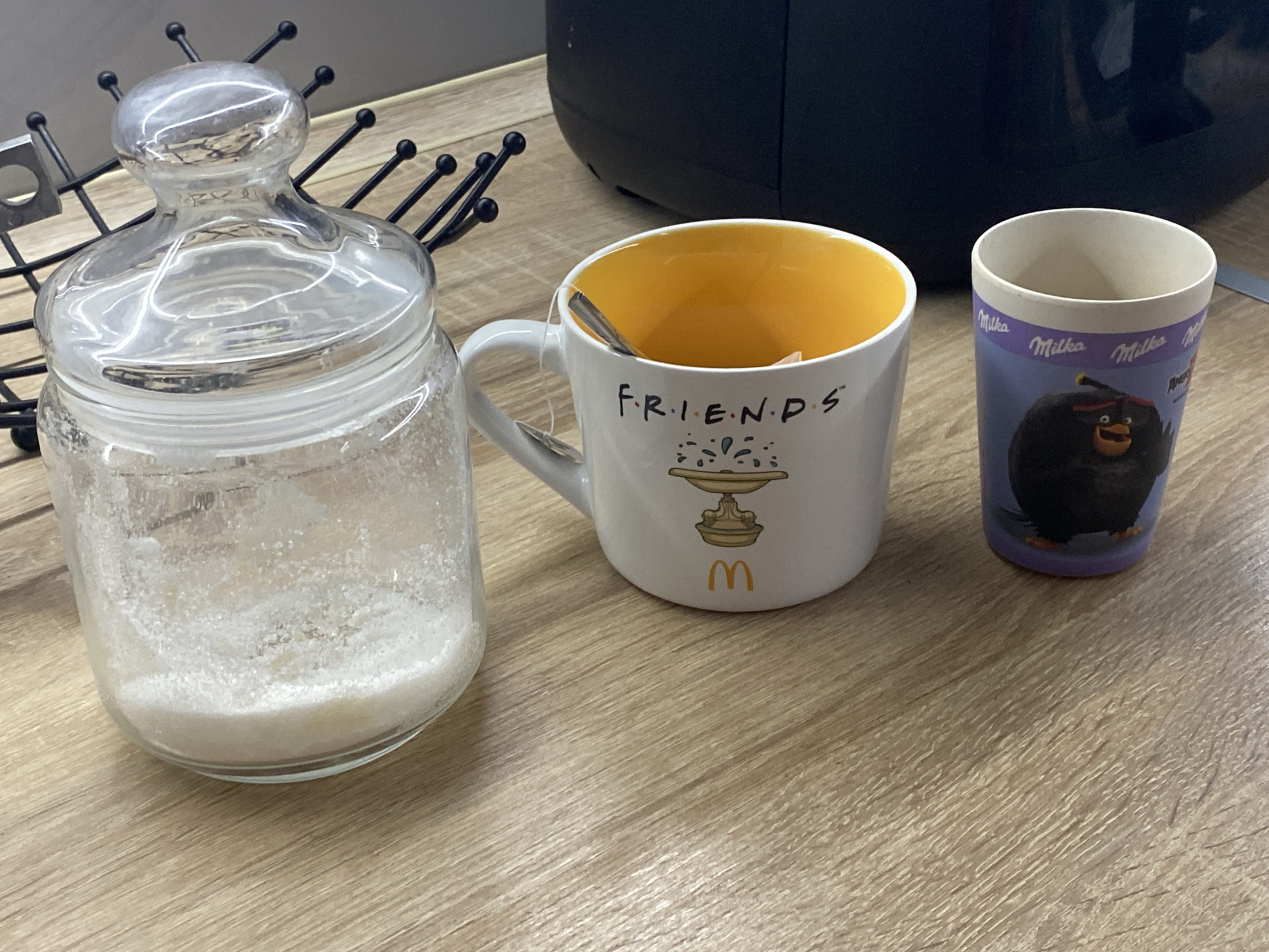A glass jar filled with a white granular substance, possibly sugar, sits on a wooden surface. To its right is a white mug with the word "FRIENDS" printed on it, and a graphic depicting a fountain with droplets. Inside the mug, a tea bag string is visible. Further to the right is a purple cup with a cartoonish black character and the word "Milka" printed on it. A black appliance is partially visible in the background. The scene appears to be indoors, on a table, with no indication of its specific location in Kryvyi Rih, Ukraine.