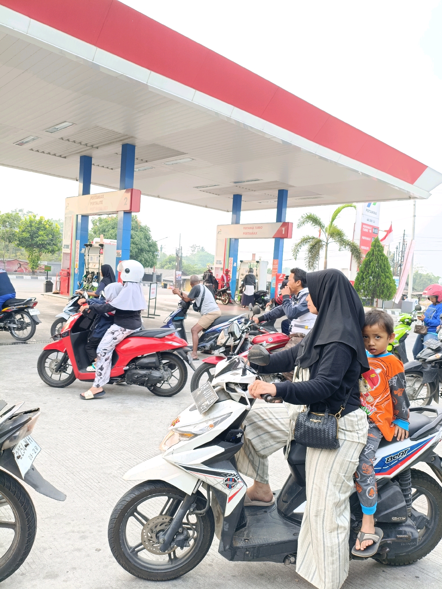 In Maguwoharjo, Indonesia, at a Pertamina gas station, a group of people on motorcycles are waiting in line. The scene is characterized by the gas station's canopy, supported by blue and white pillars, with visible signage for "PERTAMAX TURBO" and "PERTAMAX." A red motorcycle is parked in the foreground to the left, while a white motorcycle is in the center, carrying a woman in a black hijab and striped pants, and a young boy wearing an orange shirt. To the right, another white motorcycle is visible, and further in the background, several other motorcycles and individuals are present. The ground is paved with concrete.