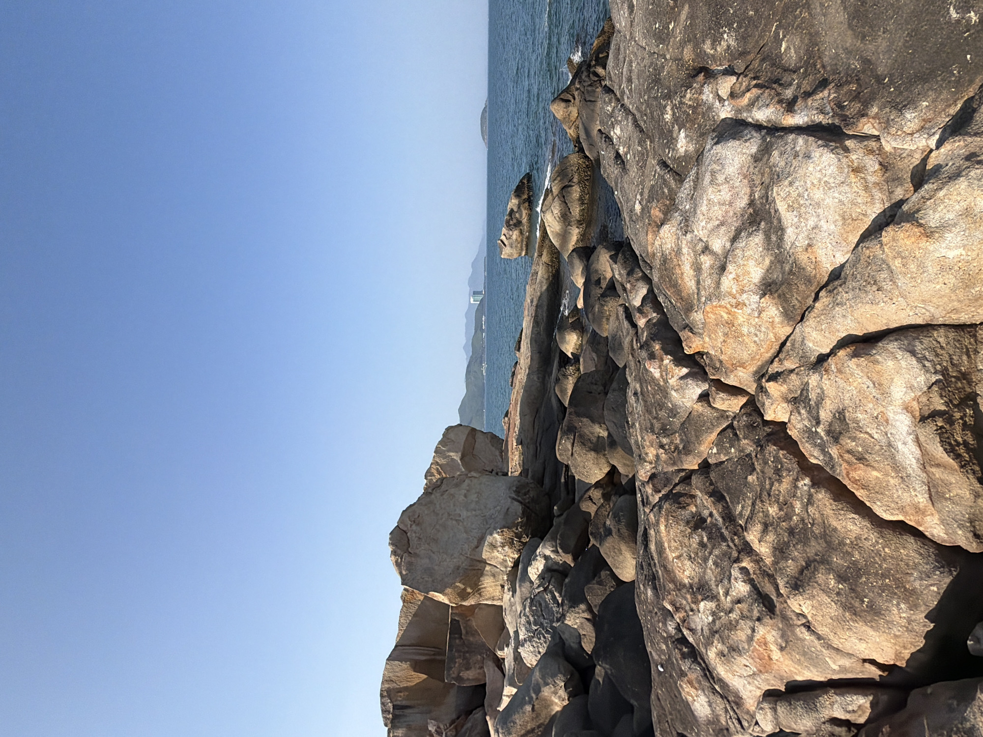 The image depicts a rocky coastline under a clear blue sky. In the foreground, large, angular rocks dominate the scene, with a textured surface displaying shades of brown, tan, and gray. These rocks appear to be part of a natural formation along the shore.
Beyond the immediate rocks, the sea stretches out, a deep blue expanse. A few smaller, rounded rocks are visible in the water, some emerging above the surface. In the middle distance, a thin strip of land with a few buildings, possibly including a hotel, is visible on the horizon, suggesting a coastal city or town. Further out, a small, green island can be seen to the right. The water exhibits gentle ripples, indicating calm conditions. The overall scene is one of a natural coastal landscape bathed in sunlight. This appears to be a section of coastline in Nha Trang, Vietnam, known for its beaches and rocky outcrops. No people or animals are visible in the frame.