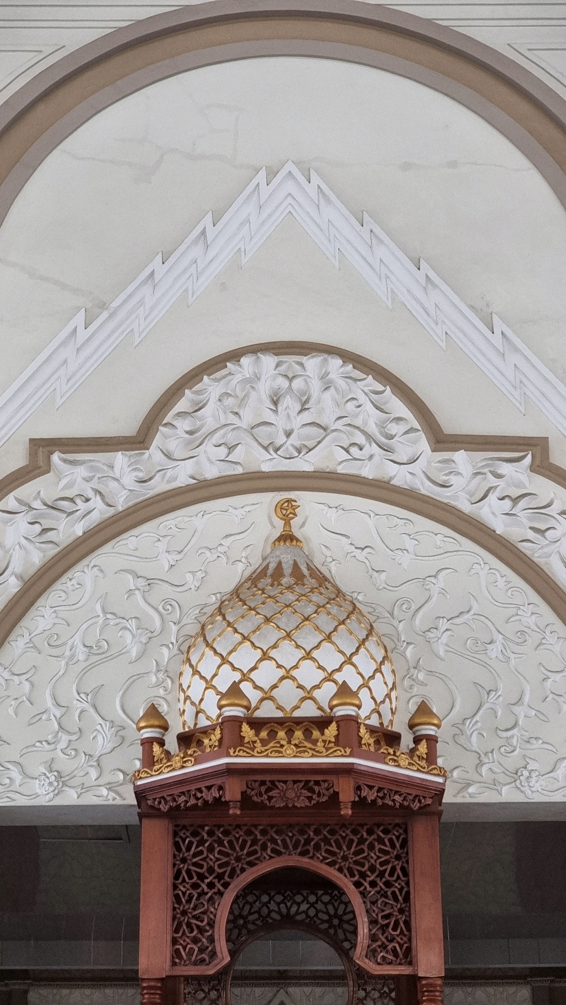 The image captures an ornate architectural detail, likely part of a mosque in Lhokseumawe, Indonesia. The central focus is a small, gilded dome with a crescent moon and star finial. The dome is adorned with a repeating pattern of golden latticework over a white base. It sits atop a decorative wooden structure, a reddish-brown hue, featuring intricate geometric carvings. Above the dome, a curved archway frames a white ceiling with a textured finish and decorative relief carvings of floral motifs. A prominent zigzag pattern in white relief is also visible on the ceiling. The overall impression is one of elaborate craftsmanship and Islamic architectural style. No people or actions are depicted.