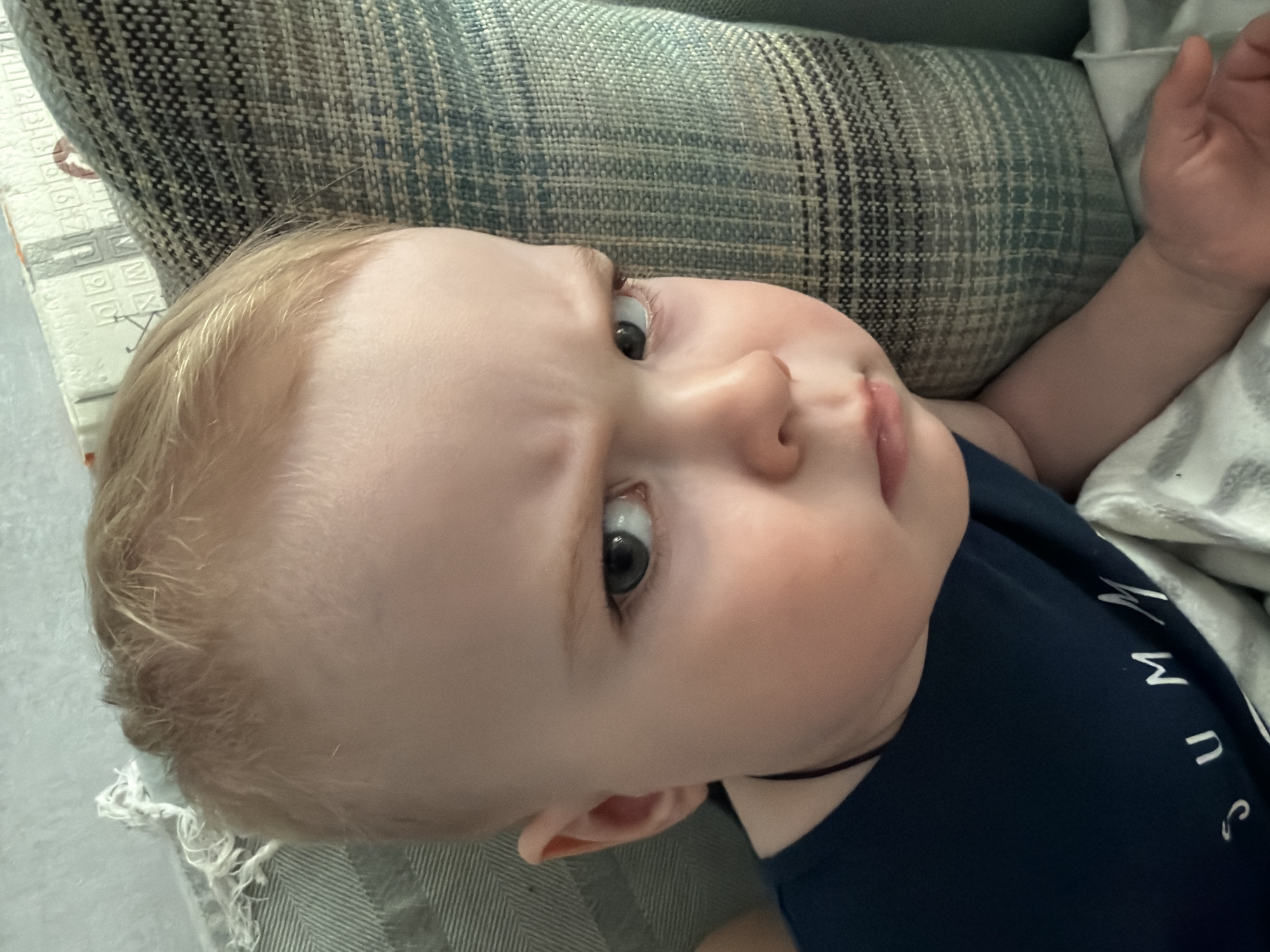 An overhead, slightly rotated close-up photograph captures a fair-skinned baby with light blonde hair and striking blue-grey eyes, resting indoors. The baby's head is positioned towards the left of the frame, gazing directly at the viewer with a curious or thoughtful expression, marked by subtly furrowed brows. The child wears a dark blue t-shirt; white letters are visible on the chest, forming "SUMM" which appears to be part of the word "SUMMER".
The baby lies next to a textured, woven pillow or cushion, featuring a blue and grey patterned fabric, which dominates the background. To the right, a white blanket with a faint pattern is partially visible, near the baby's left arm and hand. On the far left, a light-colored rug or mat shows faint, square-like patterns, possibly indicating letters or numbers. A single, fine strand of hair is visible on the patterned cushion near the baby's head. The scene suggests a well-lit, daytime setting within a home in Votkinsk, Russia.