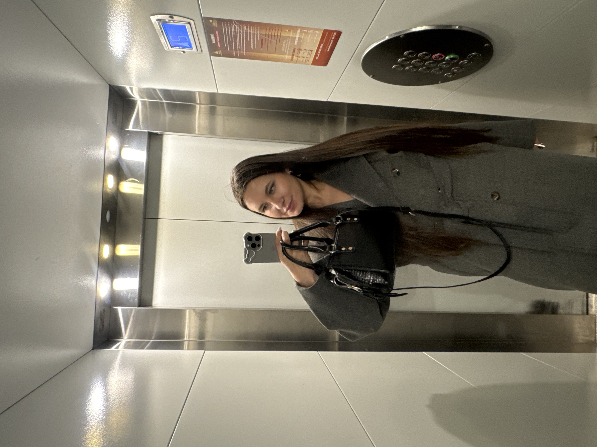 A woman stands in an elevator taking a selfie. She is wearing a gray coat and holding a black purse. The elevator has brushed metal walls with lighting strips and a floor tiled with white panels. Elevator controls and signage are visible above her head. The location is unknown and there is no information in the image to determine the city of Odintsovo or Russia.