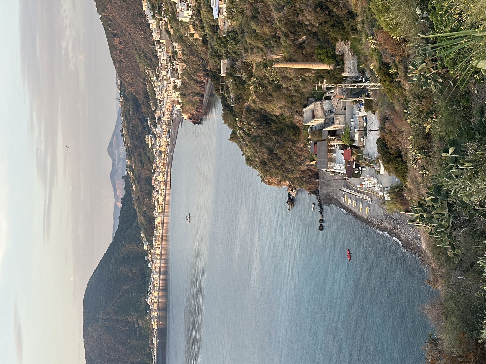 The image, presented as a landscape view, captures a serene coastal scene, likely in Lipari, Italy. The foreground on the right features a rugged hillside densely covered in green Mediterranean scrub and prominent prickly pear cacti. This slopes down to a pebbled beach, where rows of white and yellow sun loungers and umbrellas are set up, indicating a leisure area. A red kayak or canoe is visible in the calm, blue-green sea near the shore, suggesting recreational activity.
The midground is dominated by the expansive, tranquil sea. Further left along the coast, a town unfolds, characterized by numerous white and light-colored buildings densely packed along the waterfront and climbing the lower slopes of a verdant hill. A small white sailboat or catamaran is anchored or moving gently in the mid-distance on the water. On the right-hand hillside above the beach, some older, possibly industrial or infrastructural structures are visible, including a tall, rust-colored pipe or chimney and scaffolding-like supports.
In the background, beyond the town and across the bay, distant mountains or hills rise faintly under a soft, hazy sky. The time of day appears to be late afternoon or early evening, with a diffused light casting soft shadows and contributing to the peaceful atmosphere. The weather is clear and calm, with no visible clouds. There is no discernible text in the image. The architectural style and natural landscape elements align well with the volcanic island environment of Lipari, Italy.