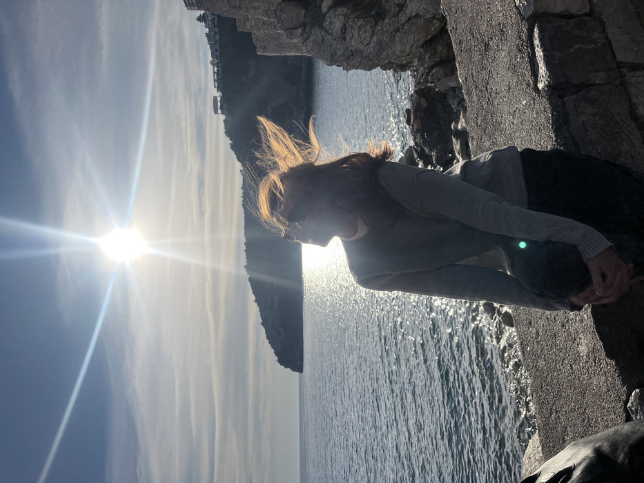 An adult female is seated on a stone-capped wall overlooking the Adriatic Sea in Budva, Montenegro, likely near the Old Town walls or a coastal promenade. She is positioned off-center to the right, facing slightly left and upwards towards the sun, which is directly behind her, creating significant lens flare and strong backlighting. Her medium-length brown hair appears windswept and is brightly illuminated, forming a halo. She wears a light-colored ribbed sweater over a high-collared white top and dark trousers, with sunglasses obscuring her eyes. Her hands are clasped in her lap. The sea occupies the left and central background, with a prominent dark, forested headland extending into the water in the middle distance. To the right, a rocky coastline with a stone wall section ascends. On a distant hill, white buildings with red roofs and other structures are visible against a partly cloudy sky with horizontal cloud streaks. The sun's glare creates a shimmering effect on the water's surface.