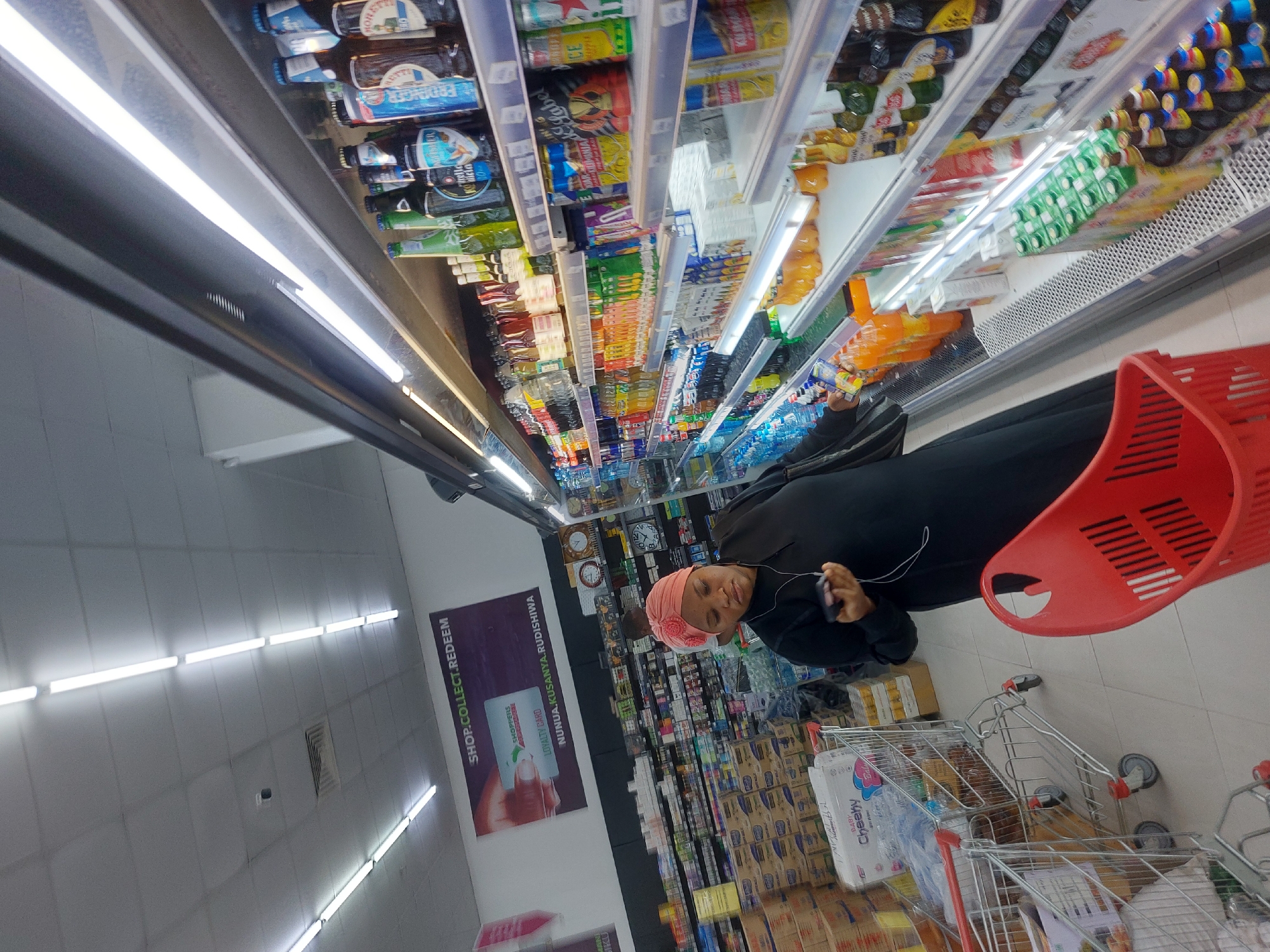 An indoor wide-angle photograph depicts a Shoppers Supermarket in Dar es-Salaam, Tanzania. The environment is a brightly lit retail space with a suspended ceiling featuring numerous fluorescent tube light fixtures and an air vent. A security camera is visible on the ceiling.
In the foreground and mid-ground, an adult female is positioned to the left of a long, multi-tiered refrigerated display unit. She is wearing a black long-sleeved garment and a pink, textured head covering. Her gaze is directed downwards at a black smartphone held in her left hand, to which white wired earbuds are connected. Her right hand holds a small, clear plastic bottle, seemingly an item selected from the refrigerated display. A red plastic shopping basket is partially visible in the lower foreground.
The refrigerated display unit, extending along the right side of the frame, is fully stocked with a diverse array of bottled and canned beverages, including various brands of soft drinks, juices, bottled water, and alcoholic beers.
To the left of the primary subject, two metal shopping carts with red handles are present. One cart contains multiple packaged items, including a prominent package labeled "Baby Cheesy Wet Wipes." Behind these carts, extensive shelving units are visible, densely packed with various consumer goods, primarily in cardboard boxes and multi-packs.
A large banner on the back wall prominently displays "SHOP.COLLECT.REDEEM" and images of a "Shoppers Supermarket Loyalty Card," with the accompanying Swahili text "NUNUA.KUSANYA.RUDISHWA." A second individual, partially obscured and wearing a light-colored top, is visible further back on the left side of the aisle. Several wall clocks are displayed on a shelf in the background to the right.