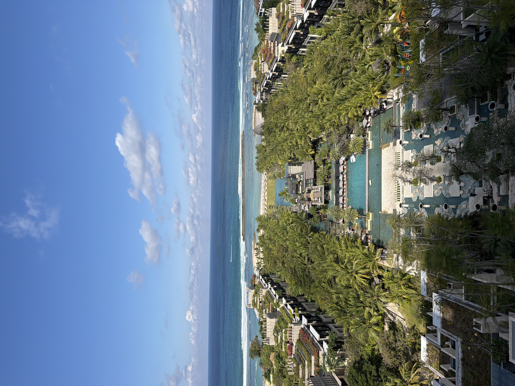 An elevated, expansive view captures a vibrant tropical coastal resort under a clear blue sky with scattered white clouds. The horizon divides the deep blue ocean from the sky, with turquoise water and breaking waves closer to the shoreline. A long breakwater extends into the ocean, and a small white boat is visible in the water.
The land is densely covered with lush green palm trees and other tropical foliage, giving way to modern resort buildings. These multi-story structures, possibly accommodations, feature balconies, some adorned with colorful flowers, and integrated rooftop greenery.
Numerous interconnected swimming pools of various shapes and sizes are a central feature of the resort area. These pools are surrounded by paved lounging areas equipped with sun loungers and umbrellas. Several individuals are visible enjoying the pools, swimming or relaxing by the water. Further inland, a children's playground with swings and a slide is nestled within a grassy area surrounded by trees.
The scene depicts a sunny day, likely late morning or early afternoon, with bright, natural light illuminating the entire landscape. The abundant tropical vegetation, resort infrastructure, and coastal setting align with the provided location context of Kuta Selatan, Indonesia, creating an atmosphere of leisure and tropical paradise. No visible text is discernible in the image.