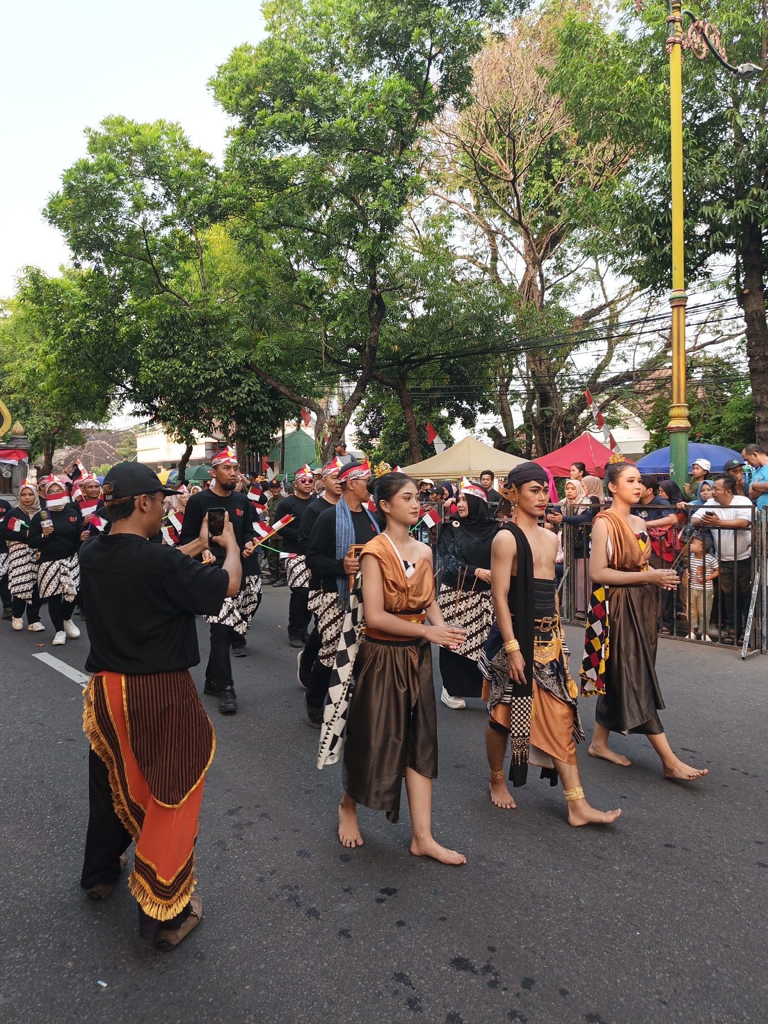 In Gayamprit, Indonesia, a cultural parade is underway on a paved street lined with lush green trees. The sky is a pale blue, suggesting it is daytime. Participants, dressed in traditional attire, walk in procession. In the foreground, a man in a black shirt and a striking orange and brown sarong-like garment is captured from the waist down, holding a smartphone to take a picture.

To his right, three individuals in traditional costumes are moving forward. The first, a woman, wears a bronze-colored draped garment with a patterned sash. The second, a man, is adorned in a black and gold top and a bronze-colored skirt-like garment, with gold armbands and anklets. The third individual, also a woman, is dressed in a similar bronze-colored draped garment.

Behind them, a larger group of people are also parading, many wearing red and white headbands and traditional patterned skirts. Some are holding small flags. Spectators are visible on the right side of the street, behind a metal barrier. A yellow lamppost stands prominently on the right. Tents and awnings are set up in the background, indicating a festive event.