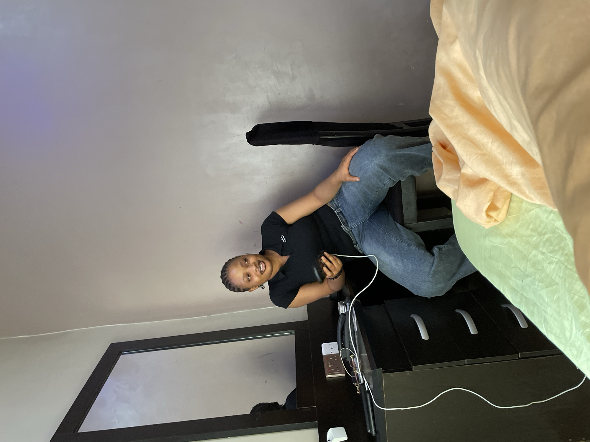 A woman with braided hair is sitting on a chair in a room. She is wearing a black polo shirt and blue jeans. The woman is holding a black smartphone in her right hand and looking to her left. A black jacket is draped over the back of the chair. To the left of the woman is a dark brown dresser with a mirror above it. Electrical outlets and a phone are visible on top of the dresser, along with a white charging cable. A light green sheet and a peach-colored blanket are visible in the foreground, suggesting a bed. The walls of the room are a neutral gray. The scene appears to be an indoor domestic setting, possibly in a bedroom or living area in Nkpor, Nigeria.