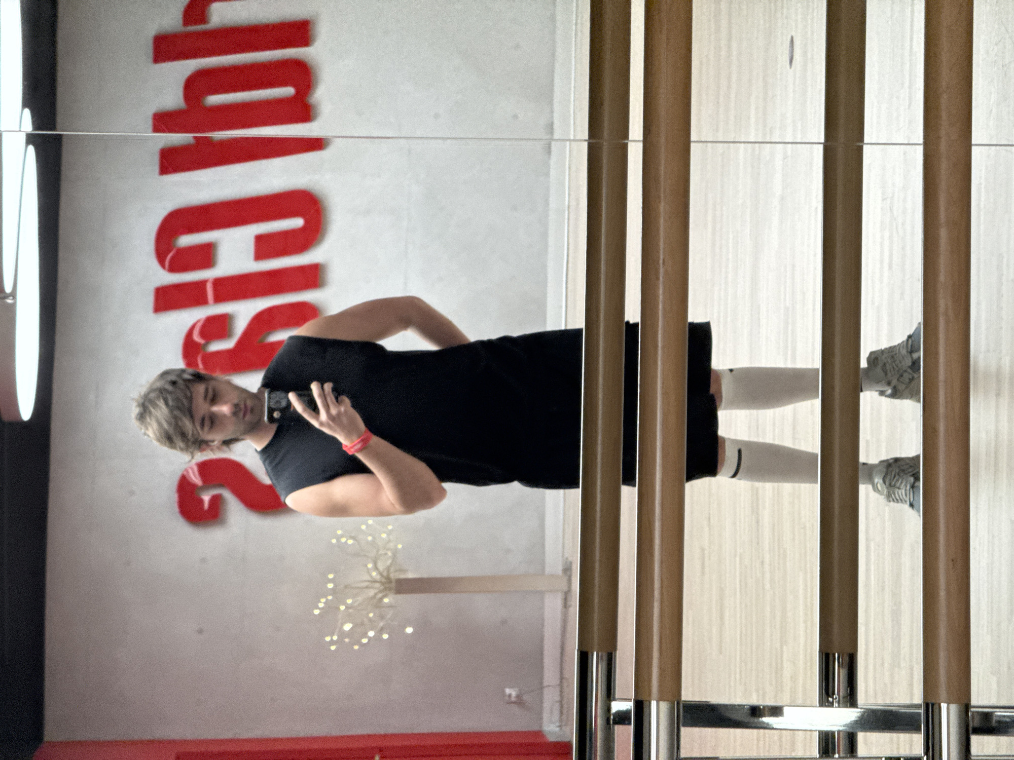 The image captures a full-length mirror reflection of a young man in a gym setting. He is wearing a black t-shirt, black shorts, white socks, and athletic shoes. A red wristband is visible on his left wrist. He is holding a smartphone in his right hand, seemingly taking the photo.
In the background, visible in the reflection, are red, stylized letters on a light-colored wall, which appear to spell out "Gym" in a large, bold font. Below the letters, a decorative lamp with small lights is visible. The gym appears to have wooden vertical elements in the foreground, suggesting a modern design. The floor is a light wood.
The image is oriented vertically, with the man appearing upside down in the reflection. The lighting suggests it could be daytime. There are no specific weather cues. The overall mood seems casual and focused on fitness. The location is identified as Moscow, Russia.