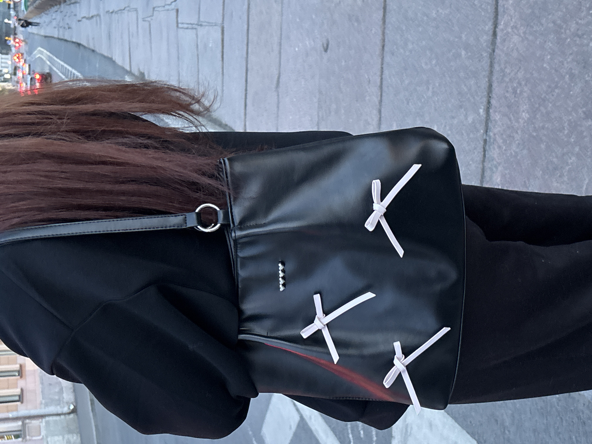 A person with long brown hair and wearing a black jacket and black pants is standing on a sidewalk in Saint Petersburg, Russia. The sidewalk is made of gray cobblestone. The person is carrying a black bag with white ribbons tied on it. There are buildings and traffic in the background.