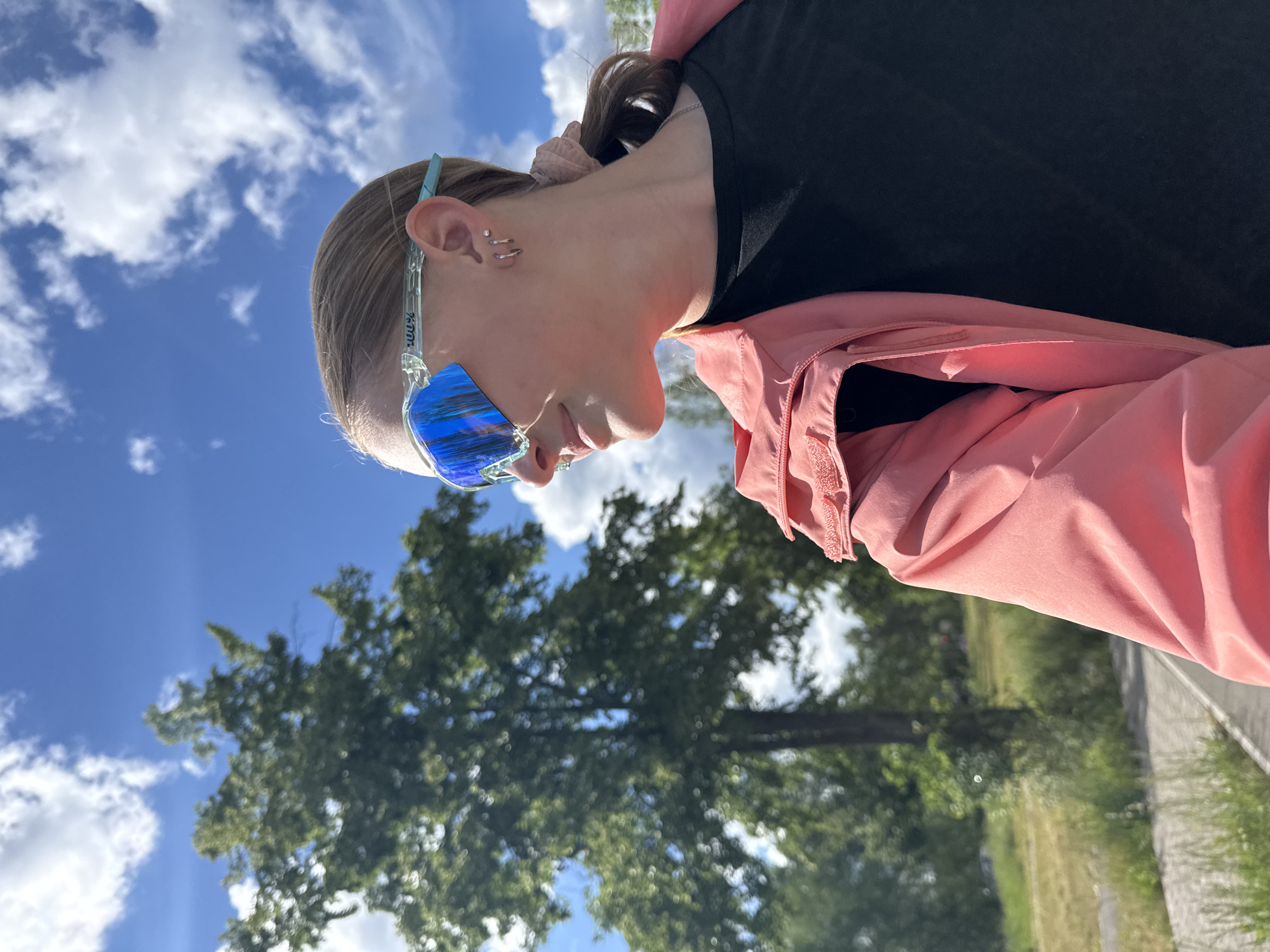 The image shows a person wearing sunglasses, a black top, and a pink jacket. The background includes a blue sky with clouds, a tree, and a path. The location is possibly Chaikovsky, Russia.