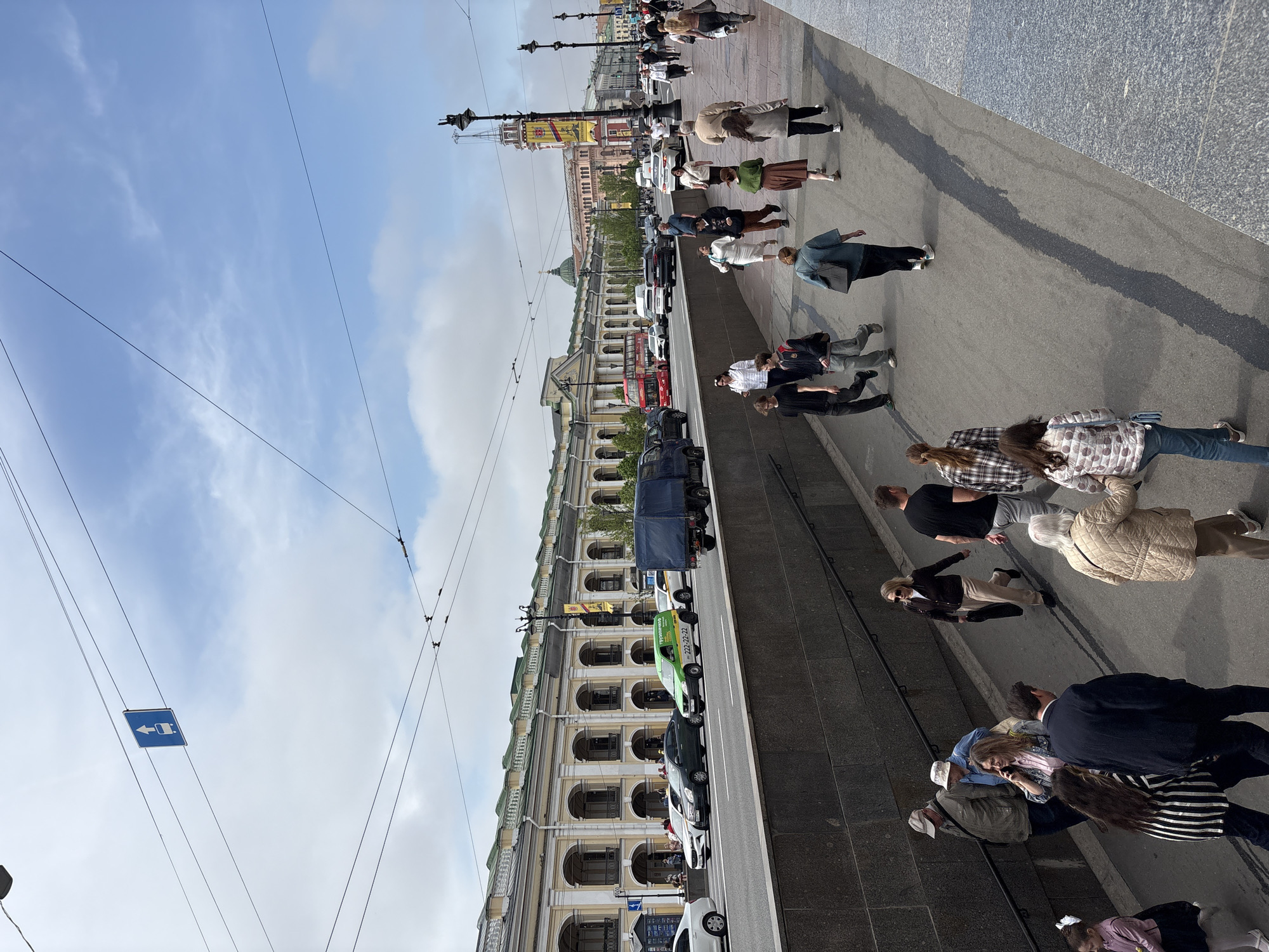 The image captures a street scene in Saint Petersburg, Russia. The perspective is a slightly elevated view, potentially from a higher building or structure. The buildings along the street have a distinctive architectural style with arched windows and detailed facades.
People are walking along the sidewalk, and cars are parked along the street. A green car with the phone number 222-22-22 on its side is visible. A blue traffic sign is seen suspended by cables in the sky. The weather appears to be clear with a blue sky with light clouds.