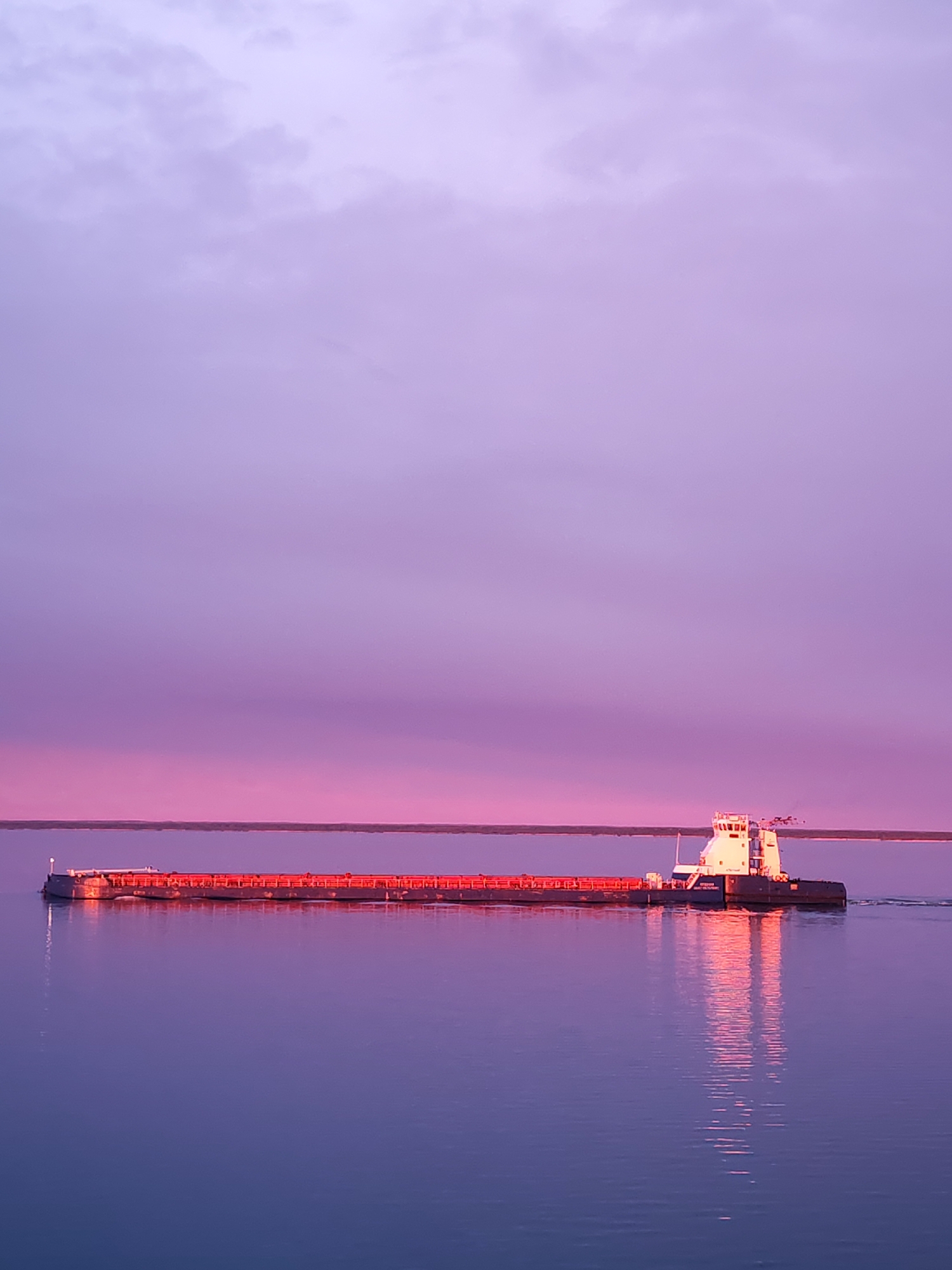 A barge travels on water under a lavender sky in Bereg, Russia. The barge, positioned horizontally in the middle of the frame, is a long, flat vessel with a white superstructure toward the right side. The deck of the barge is lined with red railing. The sky is a gradient of lavender and pink, with some faint cloud formations in the upper portion. The water, reflecting the sky, is calm and purple. A thin strip of land is visible in the distance, separating the sky and water.