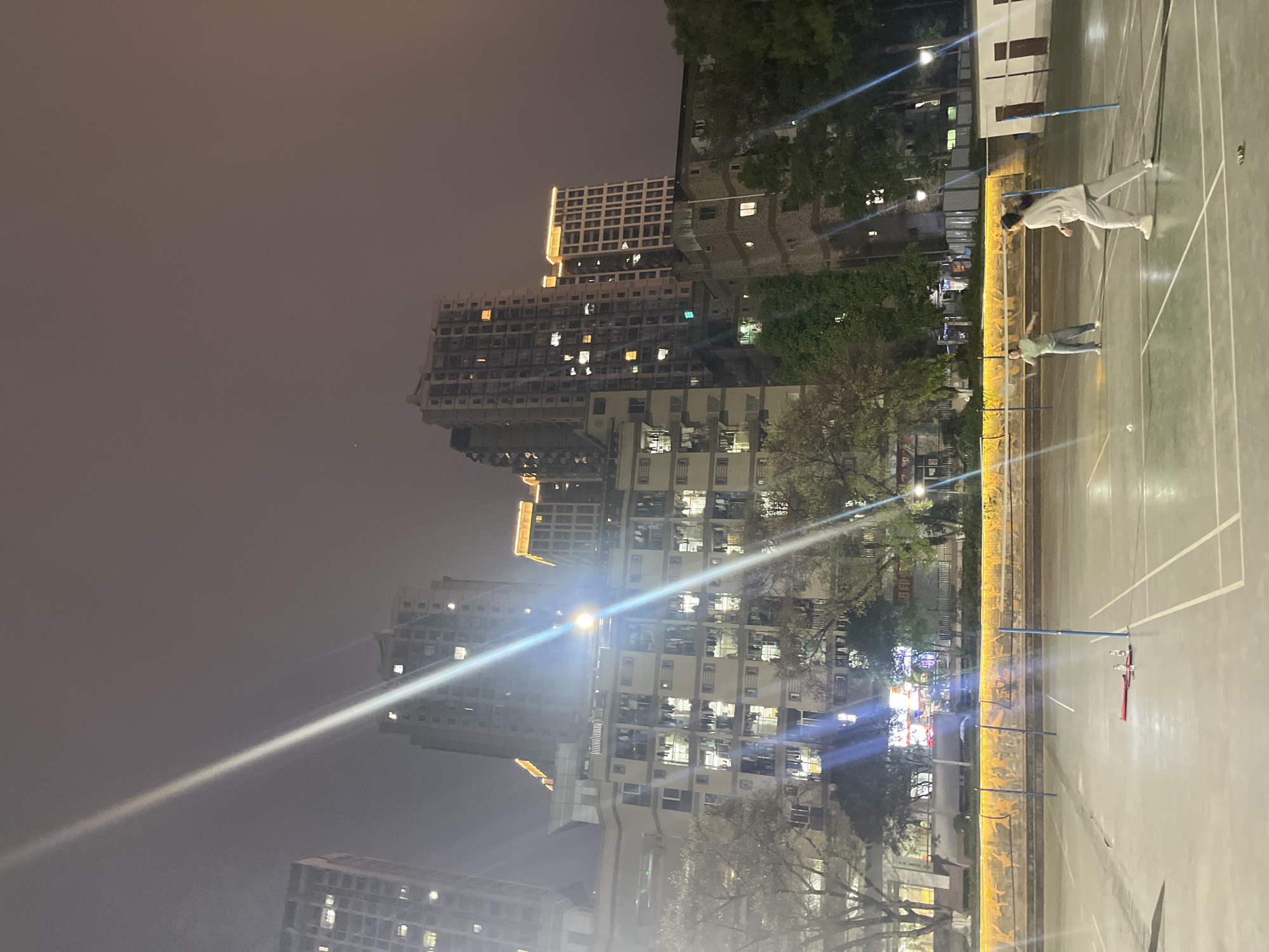 The image shows a badminton court scene in the evening with illuminated buildings in the background, likely located in Yuzhong District, China. Two individuals are on the court, one appearing to be in the middle of a badminton stroke. The buildings in the background are tall and lit up, suggesting an urban setting. Badminton equipment such as nets and rackets are visible on the court.