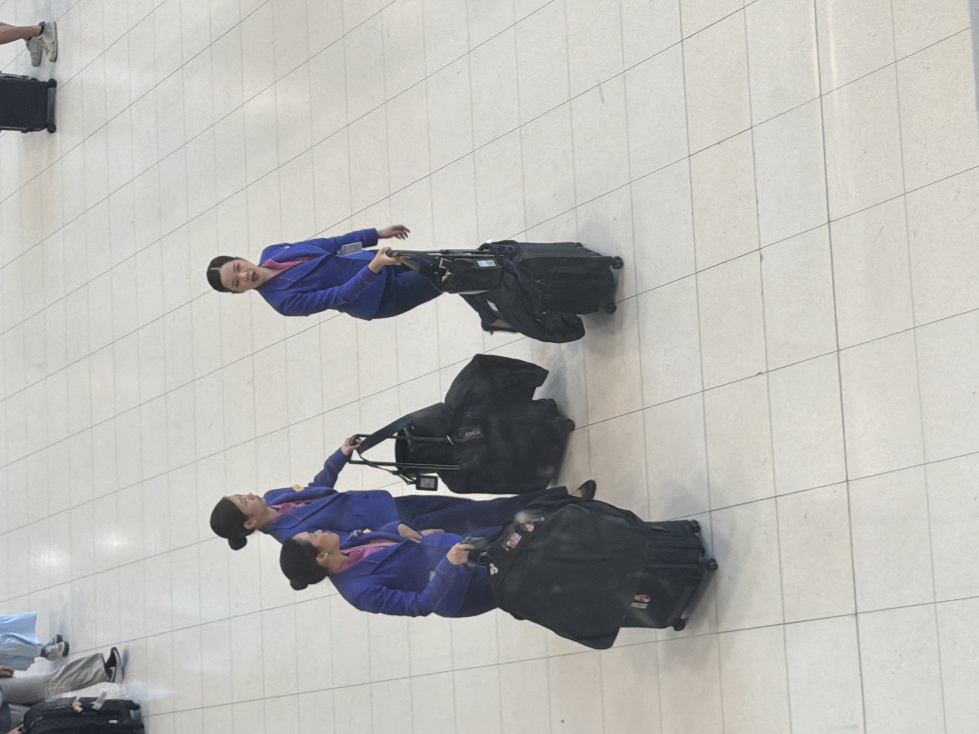 In a bright, tiled indoor environment, possibly an airport terminal in Nong Prue Subdistrict, Thailand, three individuals dressed in matching blue uniforms with pink accents are seen. The two individuals in the foreground are women, each pulling a large black wheeled suitcase. One woman, on the left, wears her dark hair in a bun and has a serious expression. The other woman, slightly behind her and to the right, also has her dark hair in a bun and is looking to her right. A third woman, further to the right and in the background, also wears the same uniform and pulls a black wheeled suitcase. Her hair is styled in a bun and she is looking forward. The floor is tiled with light-colored square tiles.