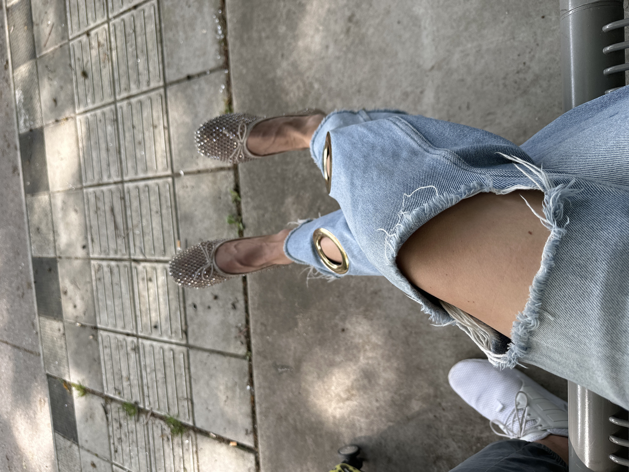 The image shows a person sitting on a bench in Dronten, Netherlands. The person is wearing ripped jeans with gold eyelet embellishments and is also wearing two different types of shoes; a sparkling flat shoe and a white sneaker. The background consists of pavement.