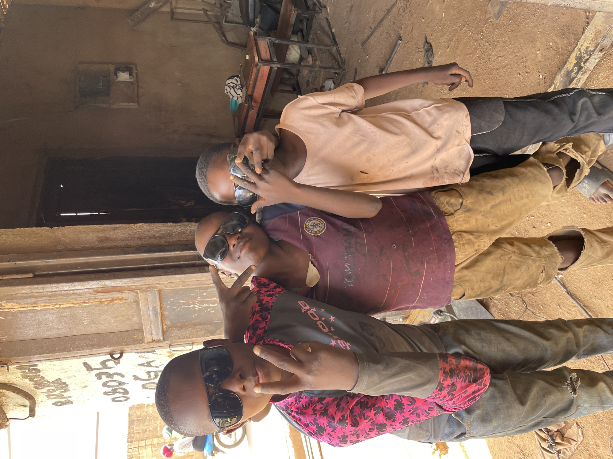 Three young boys stand close together in what appears to be a workshop in Barakin Akawo, Nigeria. All three are wearing sunglasses and making hand gestures. The boy at the bottom wears a gray and pink shirt with star patterns and dark pants. The boy in the middle wears a dirty Chelsea soccer jersey and brown pants. The top boy wears a light-colored shirt and dark pants. The setting includes metalworking equipment, a workbench with signage, and a dusty, possibly dirt floor. The scene is illuminated by natural light.