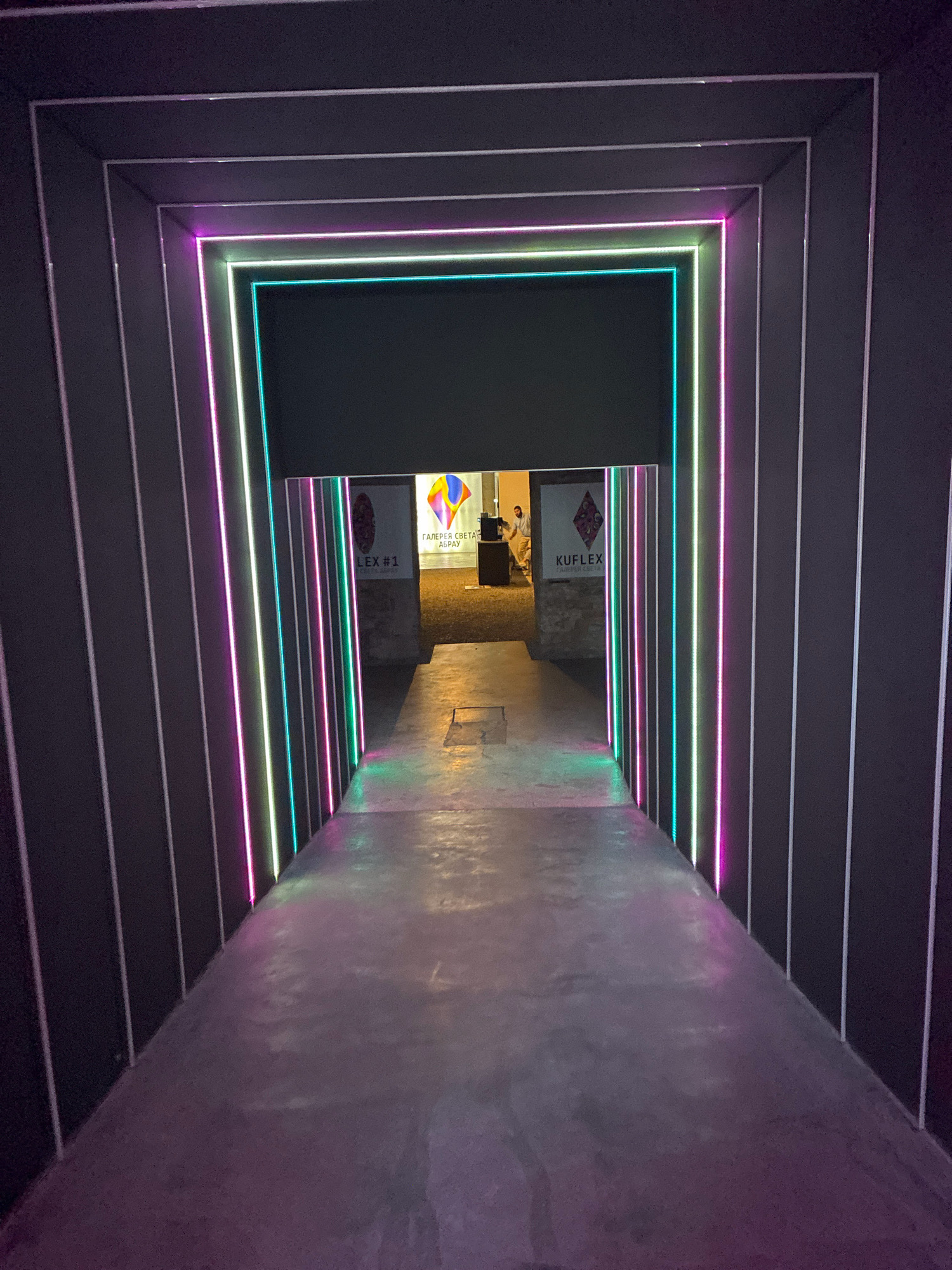The image depicts a modern architectural entrance in Abrau-Dyurso, Russia, characterized by a tunnel-like structure illuminated with vibrant neon lights. The tunnel is framed by concentric rectangular elements, each outlined with alternating purple and teal LED strips, creating a futuristic and inviting atmosphere. The floor of the tunnel is a smooth, concrete surface reflecting the colored lights. As the tunnel opens up, two doorways are visible. The left doorway features a sign with Cyrillic text and the words "LEX #1". The right doorway displays a logo with the Cyrillic text "KUFLUX" and "ГАЛЕРЕЯ СВЕТА" (Gallery of Light). A person in casual attire is partially visible standing in the background near the right doorway, suggesting activity or an event is taking place beyond the illuminated passage. The overall environment appears to be an art installation or exhibition space, given the presence of the gallery signage and the emphasis on light and design.
