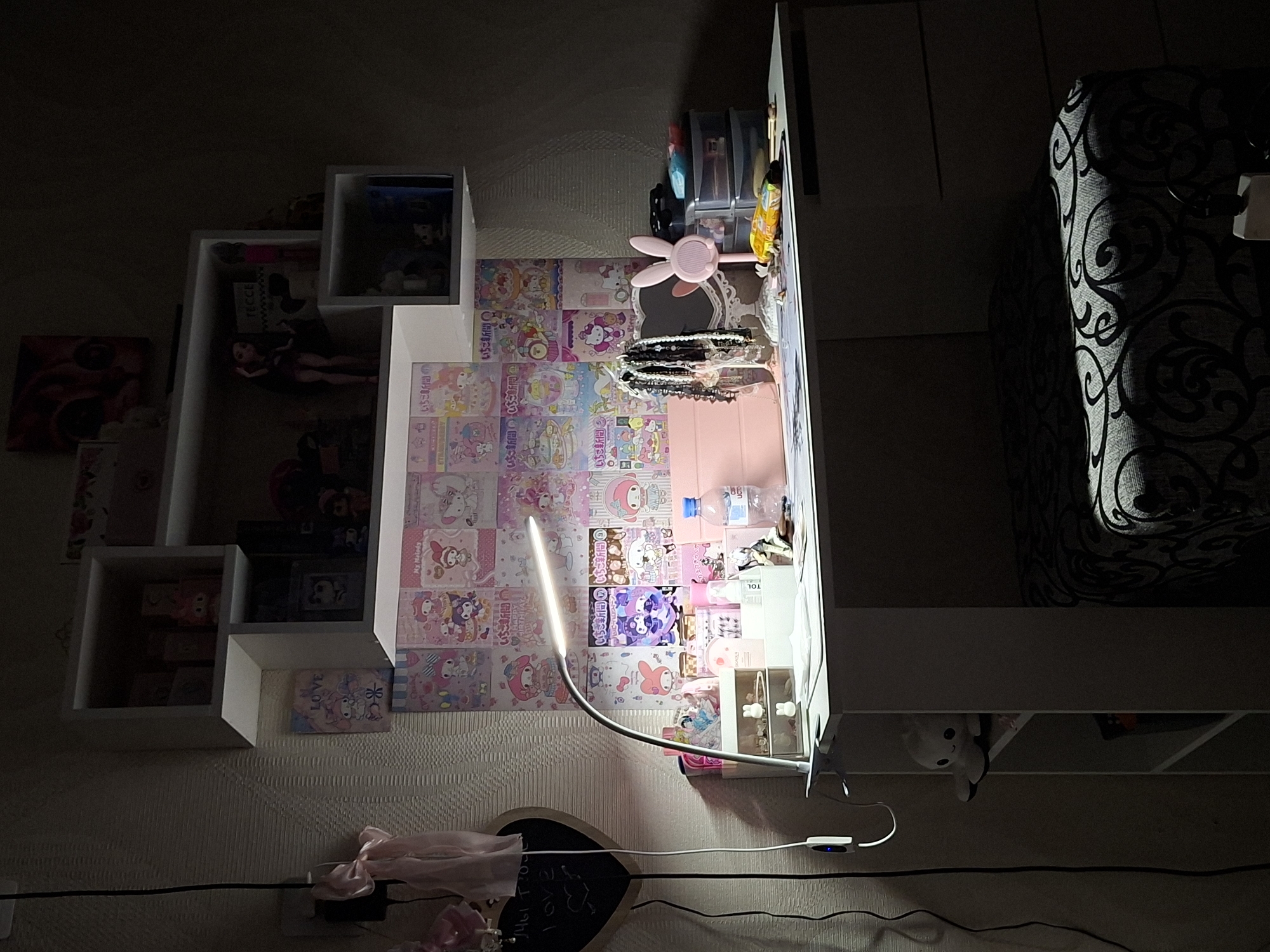 This media file captures a dimly lit interior space, likely a bedroom or personal study area in Chaikovsky, Russia, illuminated primarily by a bright, flexible LED desk lamp. The scene is presented vertically, as if looking down at a desk against a wall.
The central focus is a white desk or long shelf unit on the right, adorned with various personal items. A pink rabbit-eared mirror, possibly with a fan function, sits prominently. Nearby are multiple bracelets, a pink tablet or case, clear storage organizers holding small objects, and a water bottle. Beneath this main surface, a white stuffed animal, possibly Pikachu, peeks out from a lower shelf.
The wall behind the desk is textured and largely covered with patterned paper featuring Sanrio characters, predominantly My Melody and Hello Kitty. Several white, cubby-style shelves are mounted on the wall to the left, containing dolls, framed pictures, and other decorative items. A framed image of "LOVE" is visible on one of these shelves. Further left, a patterned artwork or poster hangs on the wall.
To the far right, a dark sofa or chair with a light-colored scroll pattern is partially visible, half-cast in shadow. At the bottom left, a black heart-shaped chalkboard displays faint white writing, with pink ribbons and wires lying around. A power strip with a blue indicator light is visible, connected to the lamp.
Visible text includes multiple instances of "いちご新聞" (Ichigo Shinbun - Strawberry Newspaper), "love," "LOVE," "HAPP" (likely part of "HAPPY"), "TOL," "TECCE," and "Мы тосе" (My tose - "We too" or "We also"). The overall impression is a personalized, decorated space with a distinct aesthetic.