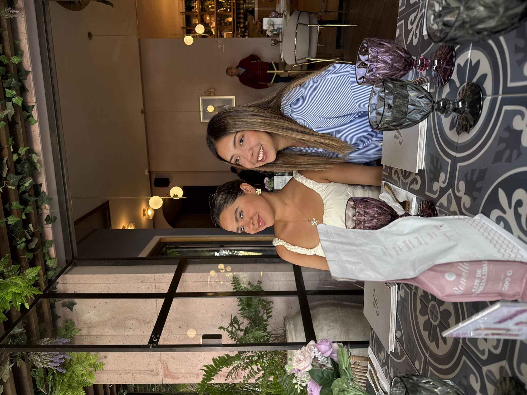 The image shows two young women seated at a table in a warmly lit, elegant restaurant or cafe, likely in Puebla, Mexico. The scene suggests an evening or late afternoon setting due to the ambient lighting from globe lights.
The woman closer to the foreground, wearing a white ruffled top, gazes subtly towards the viewer with a gentle smile. She accessorizes with a delicate necklace featuring a star-like pendant and a flower-shaped earring. Beside her, another woman in a blue and white striped shirt smiles broadly directly at the camera, her long, brown hair styled over her shoulder.
They are seated at a table covered with distinctive dark gray and white patterned tiles. The table is set with two grey ornate glasses, two purple ornate glasses, and white napkins. A pink package, labeled "Romello CAFÉ DE ESPECIALIDAD 500 G," is also visible on the table. Menus are placed beneath the napkins.
The background reveals an upscale interior with soft, warm lighting. A large glass partition or window offers a view of a stone wall adorned with lush green foliage, including ivy and ferns, and some purple decorative flowers. More potted plants, featuring ferns and purple-hued blooms, are arranged near the table. In the deeper background, a man in a red sweater is partially visible among other tables and chairs, adding to the restaurant atmosphere. The overall ambiance is inviting and pleasant.