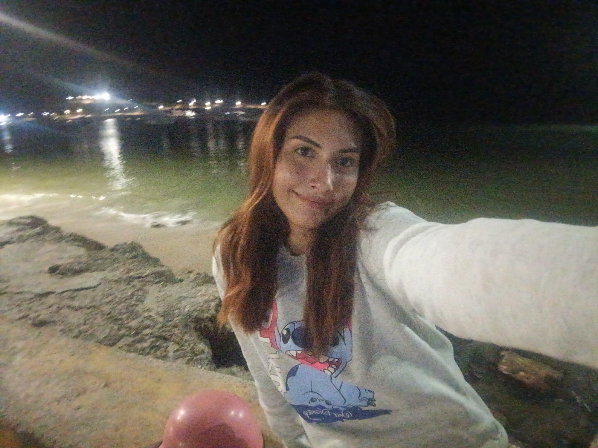 A nighttime photograph, captured in a selfie style, depicts a woman on the waterfront of Juan Griego, Venezuela. The woman, centrally positioned, has long reddish-brown hair and a light complexion, looking directly at the camera with a subtle smile. She is wearing a light grey long-sleeved sweatshirt with a graphic print of the blue Disney character Stitch, featuring pink details in its mouth. A pink spherical object, consistent with a balloon, is partially visible in the bottom left corner of the frame. The immediate foreground shows a rough, textured ground surface, likely stone or concrete, descending towards a body of water. Across this water, numerous artificial white lights from a distant settlement or structures are visible, reflecting on the dark surface of the water, which shows subtle ripples and a lighter greenish hue near the shore. The background is a dark night sky, indicating an outdoor nocturnal scene illuminated by distant city lights and possibly a nearby localized light source.