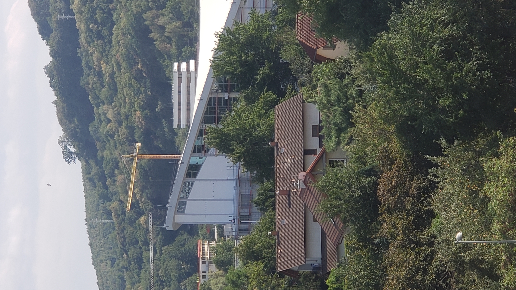 The image shows a residential area with houses in the foreground, nestled amongst trees. Behind the houses, a large modern building with a sloped roof is under construction, with scaffolding visible. Yellow construction cranes are present near the building. In the background, a tree-covered hill rises, with power lines and a transmission tower visible. The sky is partly cloudy, and a bird is flying in the distance. The scene is likely in the city of Prešov, Slovakia, given the context.