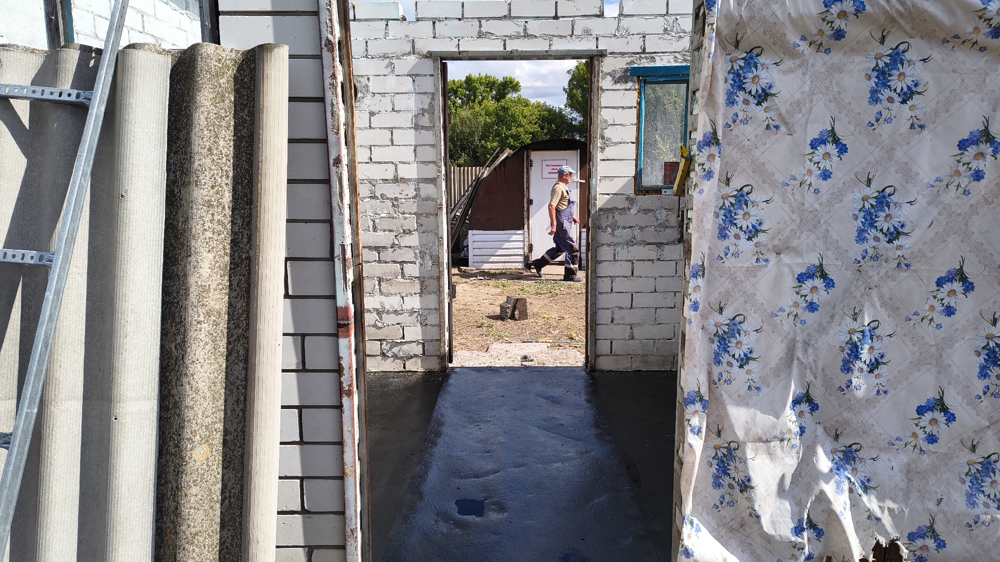 A man in blue overalls and a light-colored shirt walks through an open doorway. He is wearing a blue cap and dark shoes. The doorway is framed by a white brick structure. Inside the building, the floor appears to be newly poured concrete, with some puddles. To the left of the doorway, there are several sheets of gray corrugated material leaning against the wall, along with a metal bracket. To the right of the doorway, a light-colored fabric with a floral pattern of blue flowers and white daisies hangs down, partially obscuring the view.
Through the doorway, the scene appears to be an outdoor area with a white door in the background. Trees are visible behind the white door. The ground is dry and dusty. There is a small wooden stump in the middle of the ground. The sky is visible and appears to be partly cloudy. The time of day appears to be daytime, with sunlight casting shadows. The overall impression is one of ongoing construction or renovation, with materials stored nearby and a person working. There is text on the white door in the background, but it is too small and blurry to read clearly.