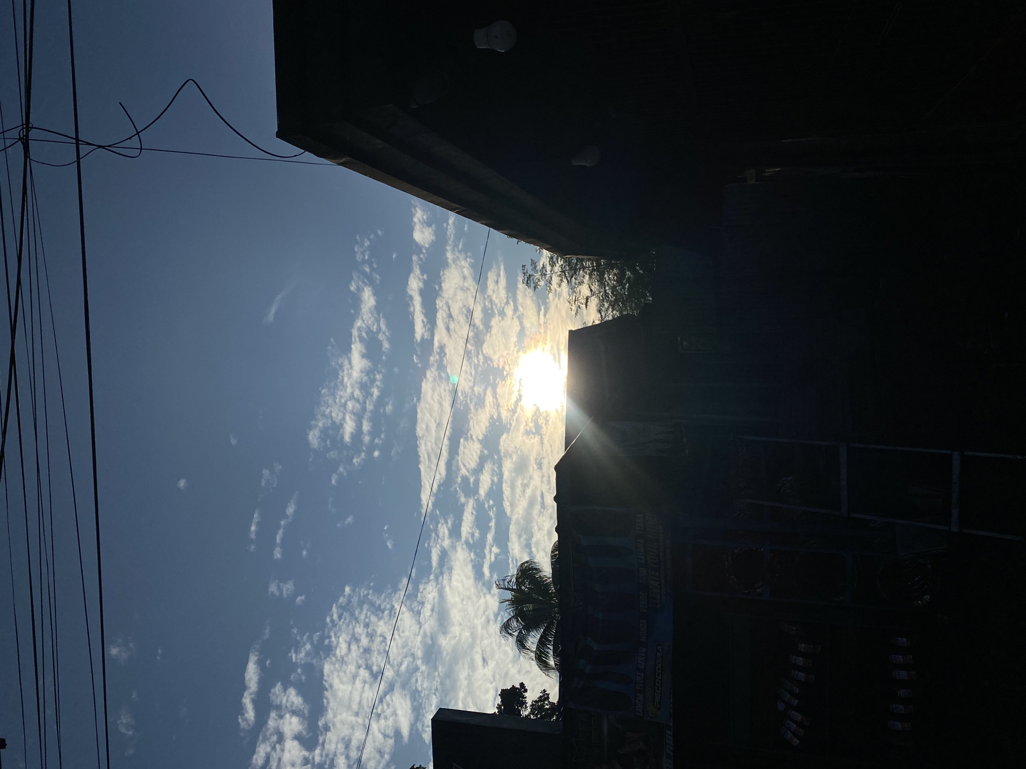 The image captures a sunset scene in the city of Davao City, Philippines. The sun is positioned low in the sky, casting a bright glare and lens flare as it peeks through scattered clouds. The sky is a vibrant blue, transitioning to warmer tones near the horizon. In the foreground, a dark, silhouetted building dominates the right side of the frame, its roofline angled upwards. Two white light bulbs are visible on the side of this building. To the left, another building, also largely in shadow, has a banner with text that is partially visible. A palm tree stands out against the sky behind this banner. Power lines crisscross the upper portion of the image, adding to the urban aesthetic. The overall impression is of a typical street view in Davao City during the late afternoon or early evening.