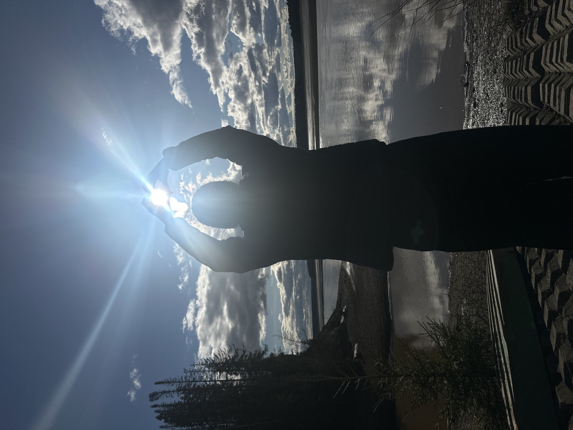 A silhouette of a person’s hands holding the sun is centered in the frame. The sun is visible as a bright light, with rays emanating outward. Behind the hands, the sky is blue with white clouds. Below the hands, there is a body of water reflecting the sky and clouds. The bottom of the frame shows trees, a wall, and some vegetation, all in silhouette. The location is likely near a body of water in Посёр, Russia.