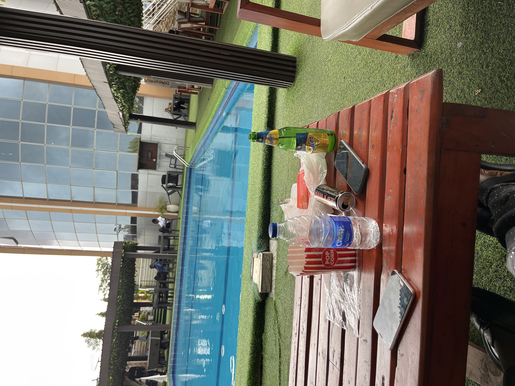 The image depicts a poolside relaxation scene at a hotel or resort in Abuja, Nigeria. A red wooden table is set up in the foreground, with a partially consumed bottle of water, a can of beer, a red and white striped popcorn box, and a mobile phone placed on it. The table is situated on artificial grass, bordering a bright blue swimming pool. In the background, a modern building with glass windows and a wooden-slatted structure are visible. Several people are seen in the distance, some near the pool and others seated at tables under a shaded area. The overall atmosphere suggests leisure and recreation.