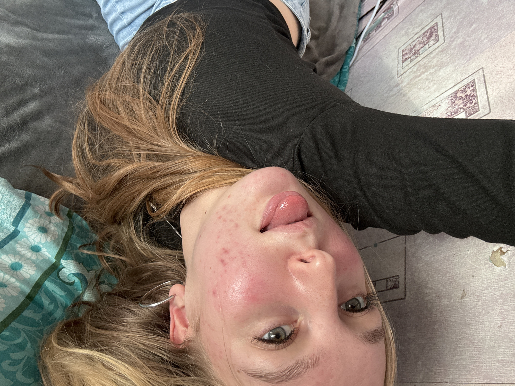 A young person, likely female, is depicted in a close-up, angled indoor photograph. She is lying down, looking directly at the camera with a playful expression, her tongue slightly extended. Her eyes are a light green or blue, and her face shows some natural redness and small blemishes. Her long, light brown hair is spread out around her head. She wears a black long-sleeved top, with light blue denim fabric, possibly jeans, visible near her shoulder. A silver hoop earring is visible on her left ear.
The setting appears to be a casual indoor space, possibly a bedroom or living room. To the left, a dark grey textured fabric, likely a blanket or couch, is prominent. Below her head, a teal and blue floral patterned fabric, possibly a pillow or another blanket, adds a decorative element. To the right of the frame, a light-colored surface with faint, darker rectangular patterns, resembling an older floor or wallpaper, forms the background. The lighting is ambient, indicating an indoor setting but providing no specific cues for the time of day or external weather. The overall mood is casual and lighthearted, suggesting a candid moment or selfie. There is no visible text in the image. The location context for this image is Perm, Russia.