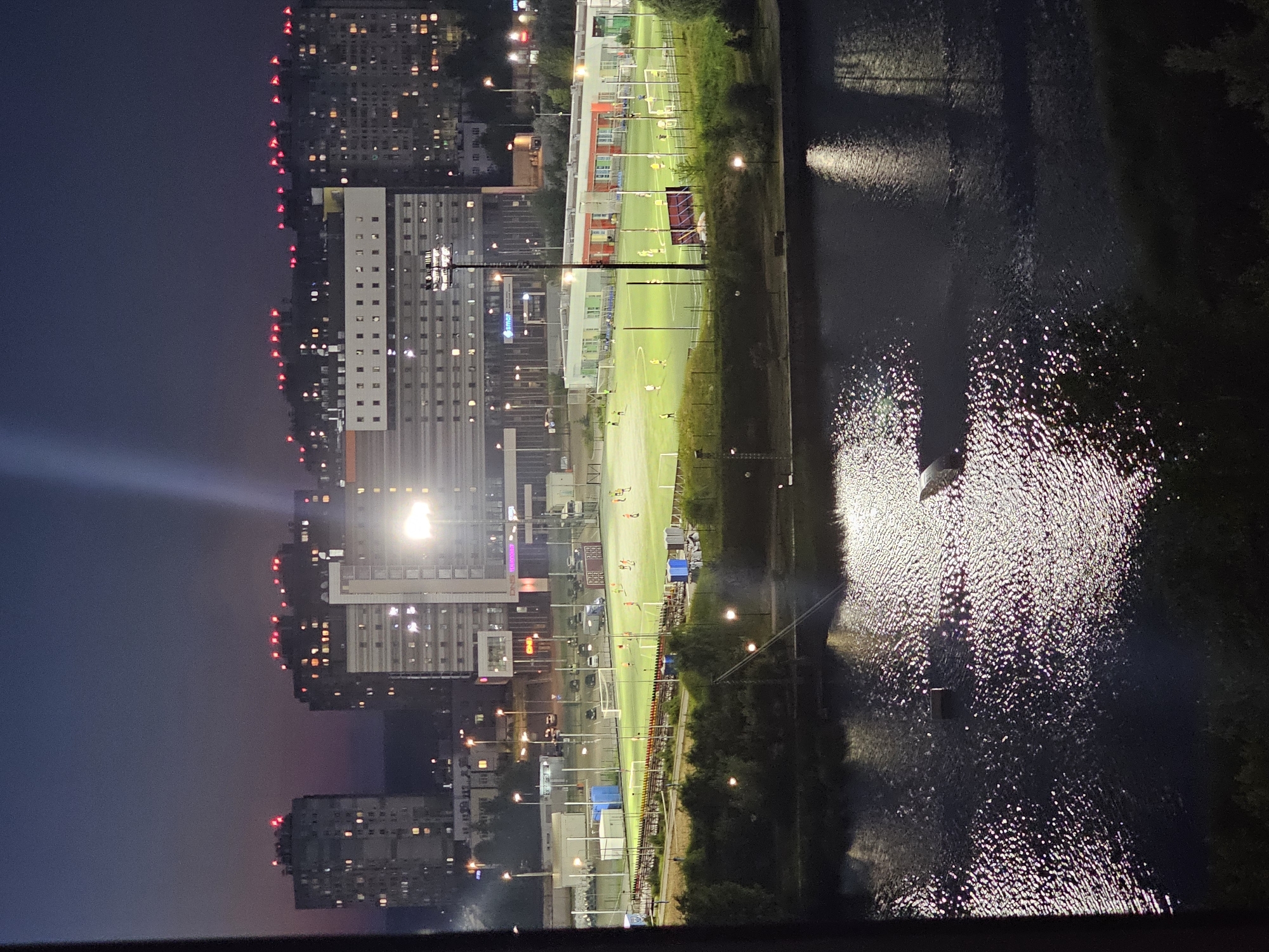 This is an aerial, nighttime photograph taken from an elevated vantage point, possibly a building, overlooking a body of water, a sports field, and several urban structures in Nizhny Novgorod, Russia. The water in the foreground reflects the lights from the surrounding city and the sports field, creating a shimmering effect on its surface. A small, dark object, possibly a boat or debris, is visible in the water.
Beyond the water, a well-lit soccer field is in operation, with several players in yellow and orange jerseys visible on the green turf. Lights illuminate the field, and a stadium-like seating area with colorful benches runs along one side. Several structures, including small buildings with orange and white facades, and portable changing rooms, are situated around the field. Parking lots with vehicles and service trucks are also present.
In the background, a complex of multi-story residential buildings dominates the skyline. These buildings feature numerous illuminated windows and red blinking lights on their rooftops, characteristic of aviation obstruction lights. The sky above is a deep twilight blue, transitioning to a lighter shade near the horizon, suggesting it is either early evening or late evening. The overall impression is of a lively urban area at night, with recreational activities taking place alongside residential and commercial development.