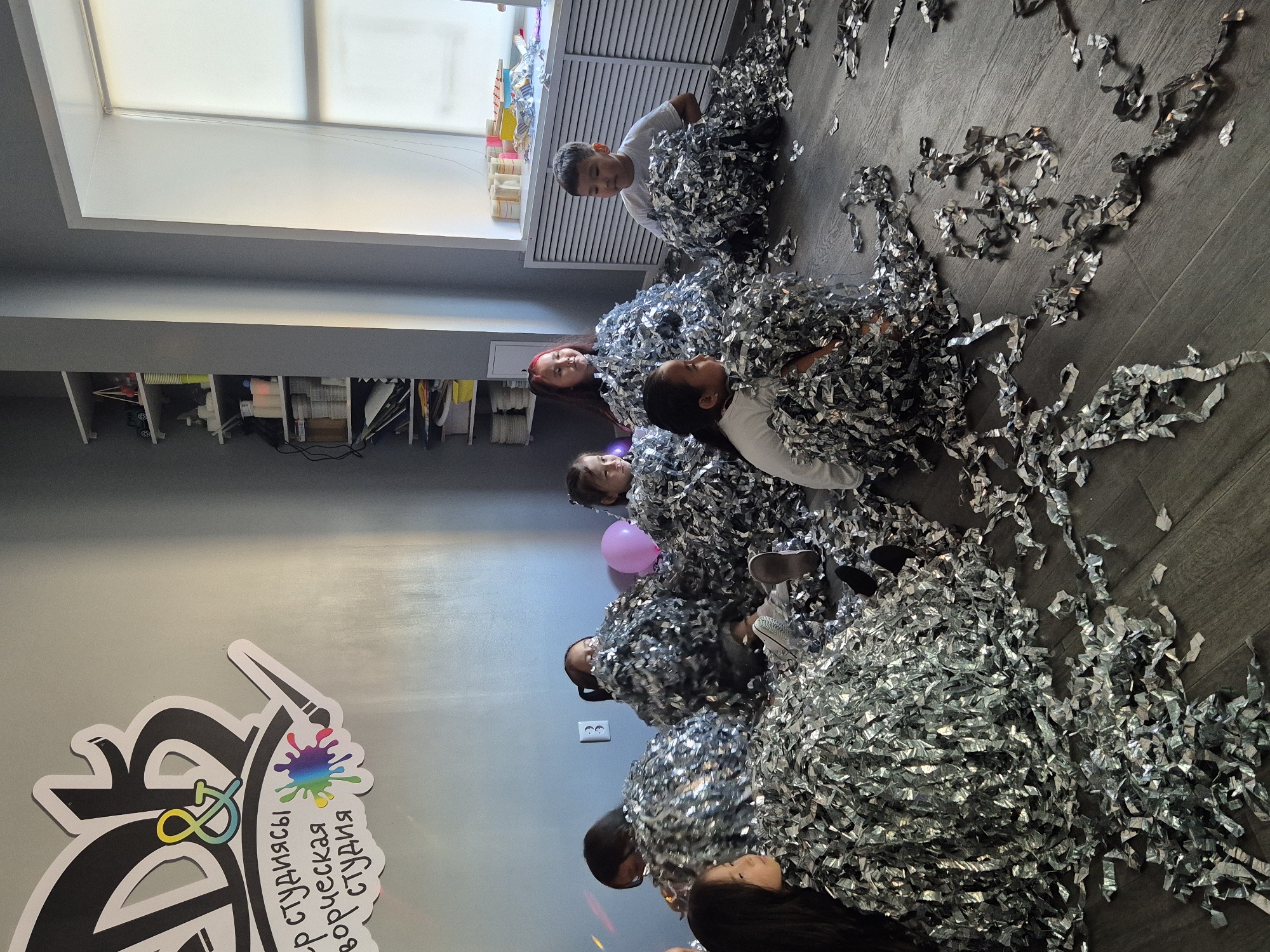 The image depicts an indoor scene, likely a children's art studio, given the logo on the wall that reads "DB & Creative Studio" in Russian. The room has grey walls and a dark wooden floor. Several children, appearing to be between the ages of 5 and 10, are sitting on the floor, each enveloped in a large pile of silver confetti. Some children are peeking out from the confetti piles. One child, a boy in a white t-shirt, is more visible and appears to be looking upwards. There is a pink balloon visible in the background, and shelves stocked with art supplies line one wall. The environment suggests a party or a fun, messy activity has recently taken place. The location is in the city of Kokshetau, Kazakhstan.