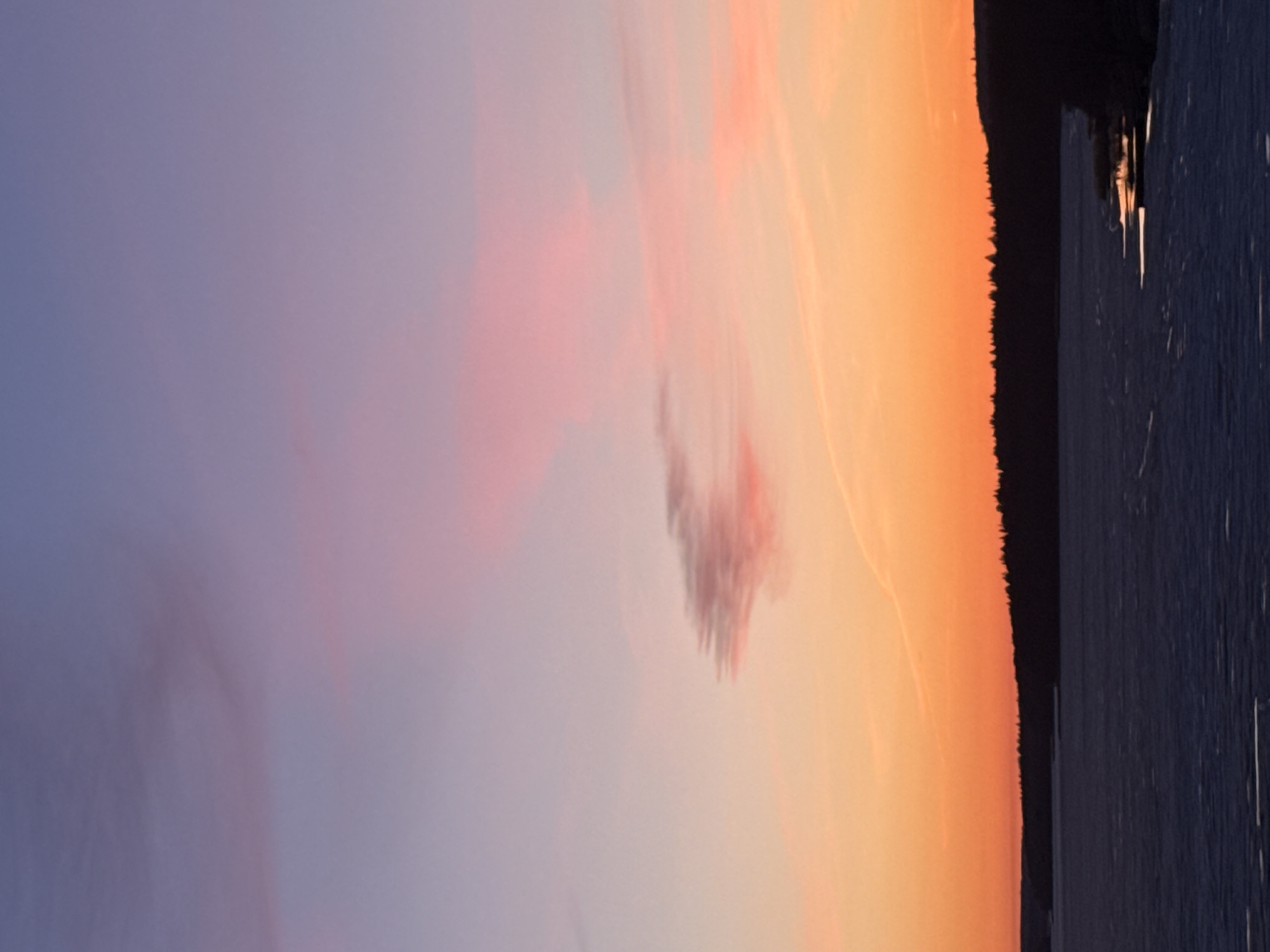 A serene landscape photograph captures a beautiful sunset or sunrise over a calm body of water in Votkinsk, Russia. The expansive sky, occupying the upper portion of the frame, showcases a dramatic gradient of colors, transitioning from a soft lavender-blue on the left to vibrant pinks and warm oranges on the right. Wispy clouds are scattered throughout, with a more prominent, reddish-brown cloud cluster noticeable towards the center-right.
A dark, jagged silhouette of a dense treeline forms the horizon, stretching horizontally across the lower middle of the image. This forested shoreline separates the colorful sky from the tranquil body of water below. The water, likely a lake or reservoir, occupies the bottom portion of the image. It appears dark and calm, subtly reflecting the ambient light from the sky, with faint streaks or ripples visible on its surface, particularly closer to the landmass.
The scene conveys a profound sense of natural beauty and peacefulness, characteristic of either dawn or dusk. There are no people, man-made objects, or visible text present in the image.