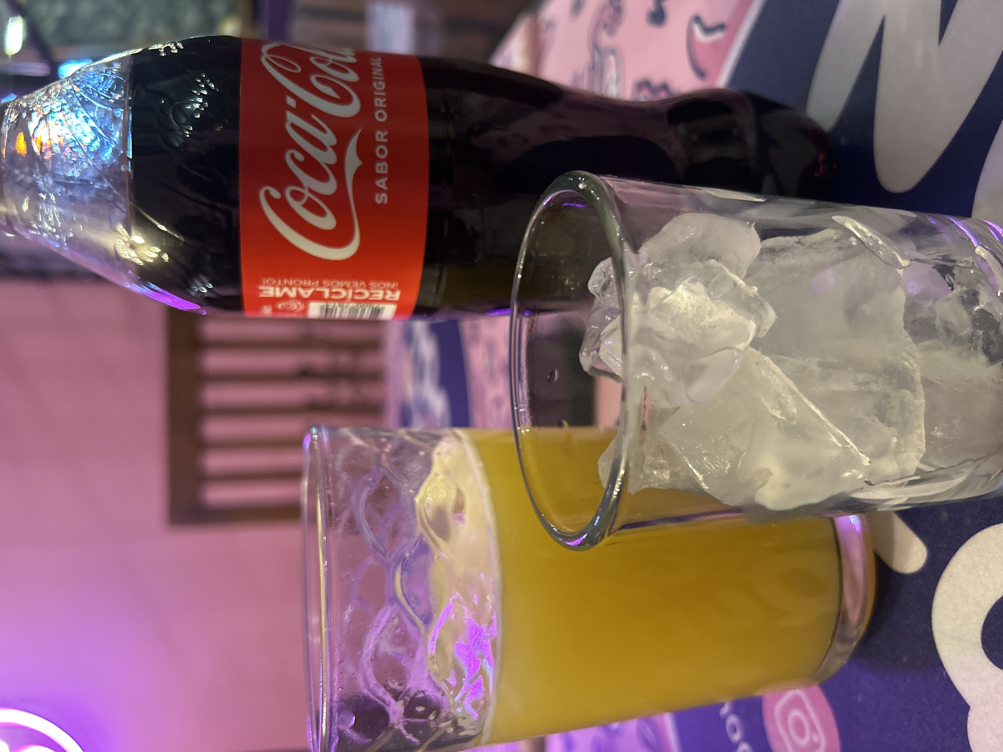 The image displays a close-up, horizontally oriented shot of beverages and glasses on a patterned surface, likely a table, in an indoor setting in Puebla, Mexico. The scene features a dark glass bottle of Coca-Cola with its iconic red label, positioned near two clear glasses.
The Coca-Cola bottle is visible from its cap down to about halfway along its body. Its label clearly displays "Coca-Cola" in white cursive lettering, with "SABOR ORIGINAL" (Original Flavor) beneath it. Further down, partially visible in the bottom left of the label, is "RECÍCLAME" (Recycle Me) in white, indicating Spanish text, consistent with the location context. The bottle's upper part shows reflective light, possibly from a light source above or nearby, creating vibrant highlights on the plastic wrap around the cap area.
Next to the Coca-Cola bottle is a tall, clear glass filled with numerous irregularly shaped ice cubes, suggesting a cold beverage is intended. Below this glass, closer to the viewer, is another clear, textured glass holding a bright yellow or orange liquid, likely orange juice or a similar citrus-based drink. The textured pattern on this glass refracts the light, creating interesting visual effects.
The background features a soft, blurry purple and pink glow, possibly from ambient lighting, neon signs, or decorative elements within the establishment. A darker, blurry rectangular shape in the mid-background might be a piece of furniture or part of the interior decor. The surface beneath the drinks appears to be a tablecloth or a similar covering, with faint patterns in shades of pink, white, and blue. The overall impression is one of a casual refreshment or dining experience, potentially an evening out given the artificial lighting. No people are visible.
