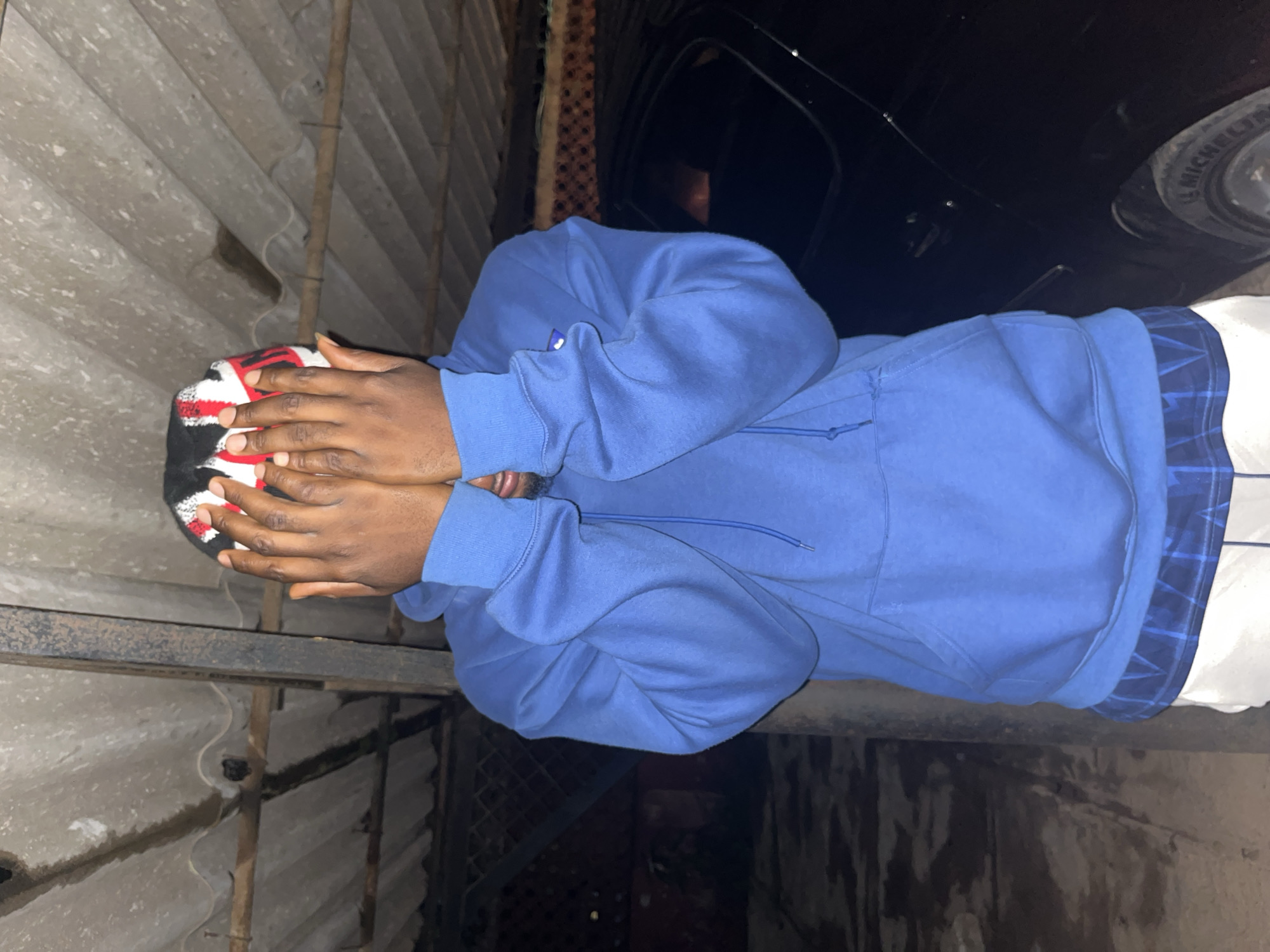 In Jos, Nigeria, a person is captured in an image, their face obscured by their hands and a beanie with red, white, and black stripes. They are wearing a bright blue hooded sweatshirt over a blue plaid shirt, with the hem of white shorts visible below. The backdrop suggests a rustic setting, with a corrugated metal roof overhead supported by wooden beams and a metal pole. A dark-colored vehicle is parked to the right of the frame, partially visible. The lighting indicates it is nighttime.