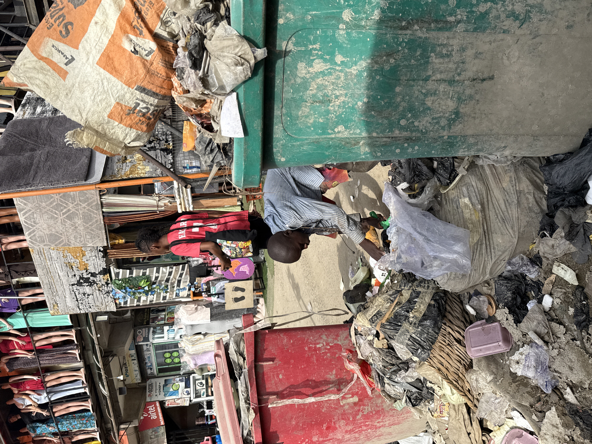 This image depicts a bustling outdoor market scene in Ado, Nigeria, under bright daylight. In the immediate foreground, a significant accumulation of waste and discarded items dominates the right side. A man with dark skin and a shaved head, wearing a blue and white striped long-sleeved shirt, is bending over this large pile of trash, appearing to sort through it. The refuse includes numerous black and clear plastic bags, various plastic debris, and other general waste. Surrounding this pile are two large, weathered bins: one green and one red.
Behind the man, the ground is dusty and unpaved. To the left, a woman with braided hair, dressed in a red t-shirt and black trousers, stands near a market stall, looking down at items displayed on a surface. This area transitions into a lively market background, filled with various informal shops and stalls.
Notable details in the market section include:
- A large, torn orange and white sack, likely a cement bag, with visible text "LAFARGE" and "SUPASET CEMENT."
- Stacks of textiles, carpets, and other household goods are arranged on shelves and surfaces.
- Further back, a display of mannequins or mannequin parts, dressed in various garments, can be seen.
- Numerous shopfronts display a wide array of products, including electronics and other merchandise.
Visible text on shop signs includes the distinctive red "Airtel" logo, along with partial names like "OZIK," "SWAP," and "jozik." A small sign featuring two black footprints is also present. The overall scene captures the vibrant, dynamic, and somewhat unkempt atmosphere of daily life and commerce in a busy urban market.